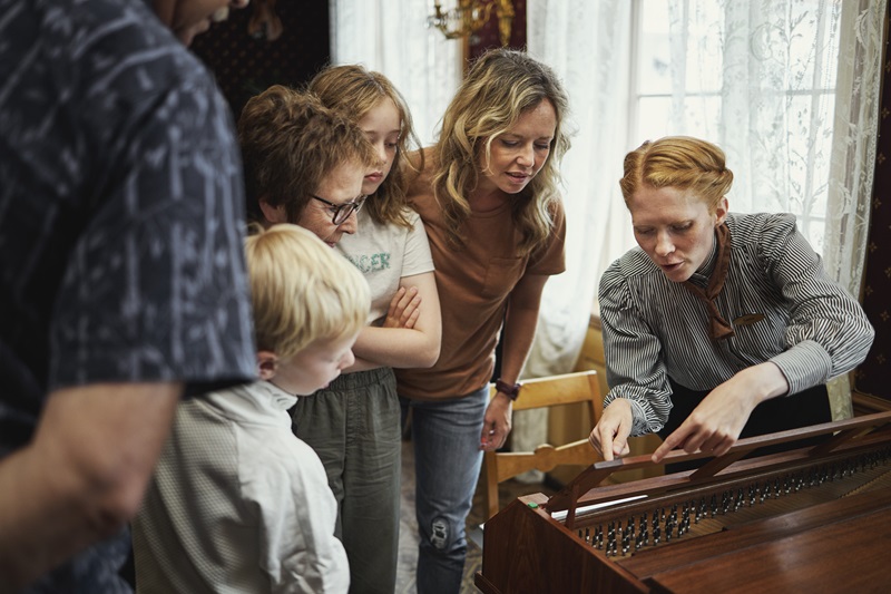 En gruppe mennesker, både barn og voksne, er samlet rundt en kvinne som demonstrerer et musikkinstrument, trolig et cembalo eller lignende klaviaturinstrument. Hun peker på de indre delene, som strenger eller mekanikk, mens de andre følger nøye med. Rommet har blonder foran vinduene og et klassisk preg, med naturlig lys som skaper en varm og engasjerende atmosfære. Bildet formidler en læringssituasjon med fokus på musikk og historisk formidling.