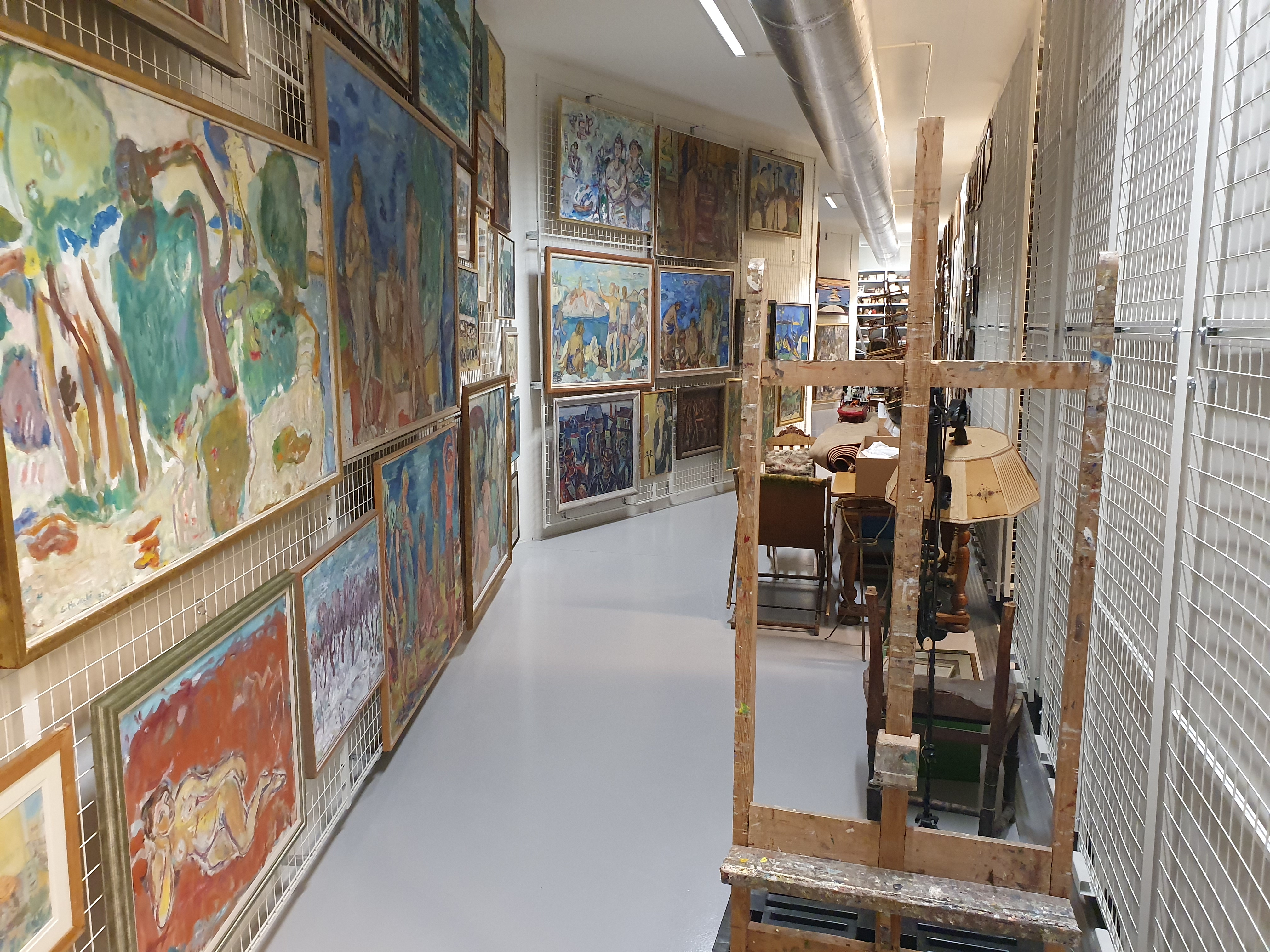 Smalt rom i et kunstlager eller galleri med fargerike malerier på begge vegger, festet til metallnetting. Bildene varierer i størrelse og stil, med mange abstrakte og impresjonistiske uttrykk. Til høyre står et stort staffeli og et bord med stoler, som antyder et arbeidsområde for kunststudier eller restaurering. Rommet er godt opplyst med taklys og fremstår ryddig og profesjonelt, trolig en del av et museum eller kunstinstitusjon.