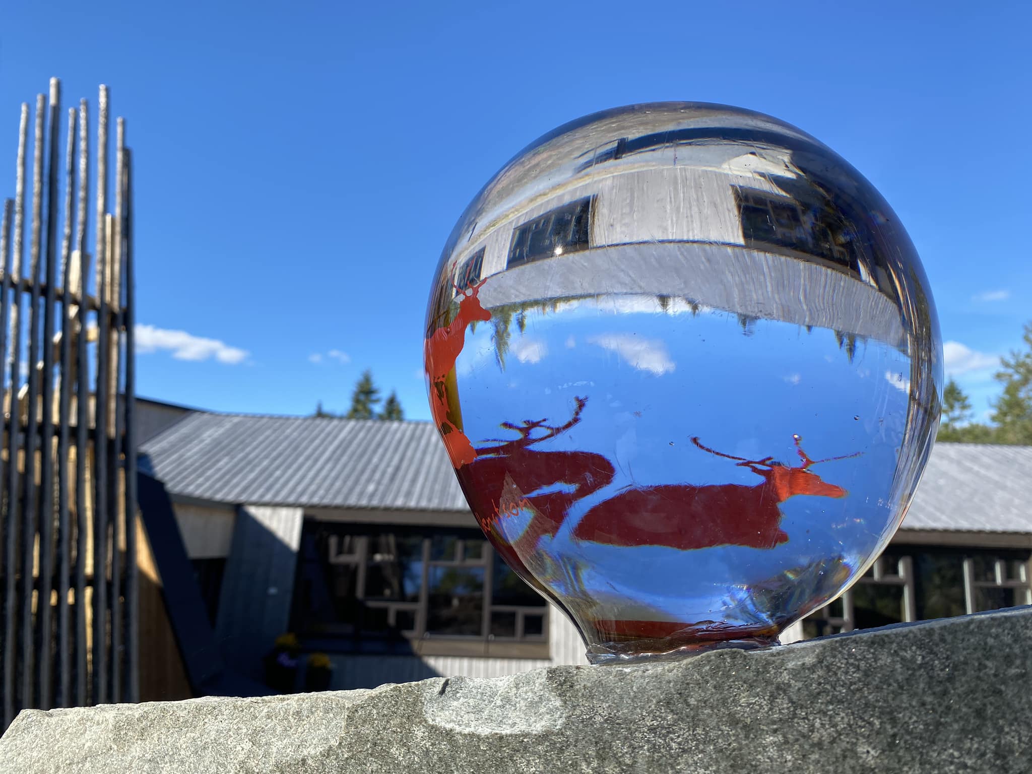 En glasskule ligger på en steinflate utendørs og reflekterer omgivelsene på en optisk forvrengt og opp-ned måte. Inne i kulen sees et bygg med grått tak, blå himmel og røde hjortefigurer. Til venstre står en høy konstruksjon av trestolper. Lyset og refleksjonene skaper et kunstnerisk og tankevekkende uttrykk, der natur, arkitektur og visuell lek med perspektiv møtes.