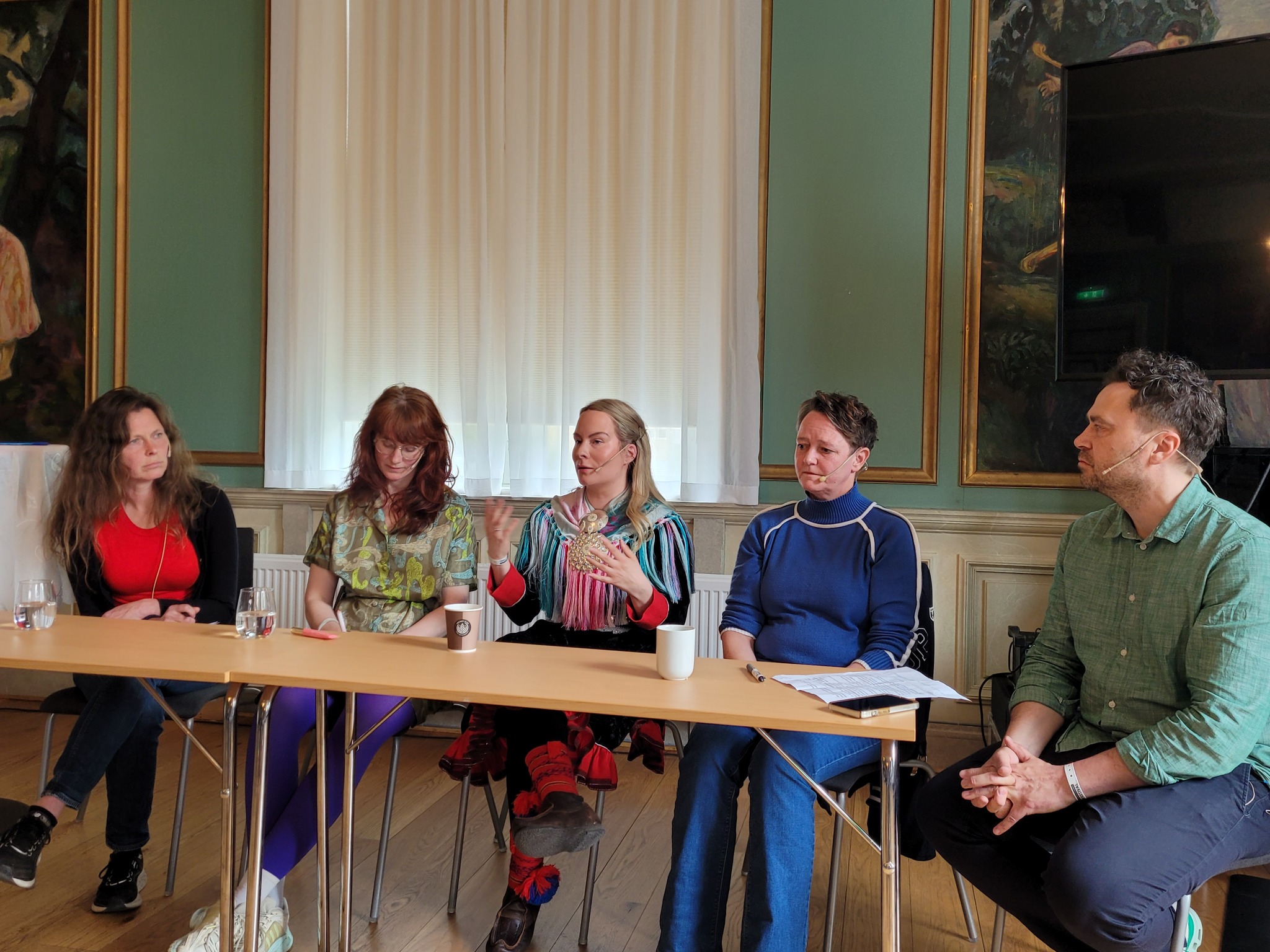 Fem personer sitter bak et bord iført klær i ulike farger. Fire kvinner og én mann helt til høyre i bildet