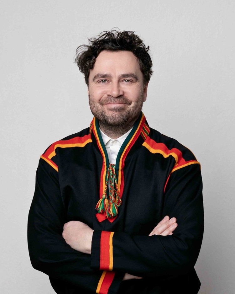 En mann står midt i bildet iført svart samisk kofte med røde og gule detaljer. Han har brunt hår, smiler mot kamera og har armene krysset foran kroppen.