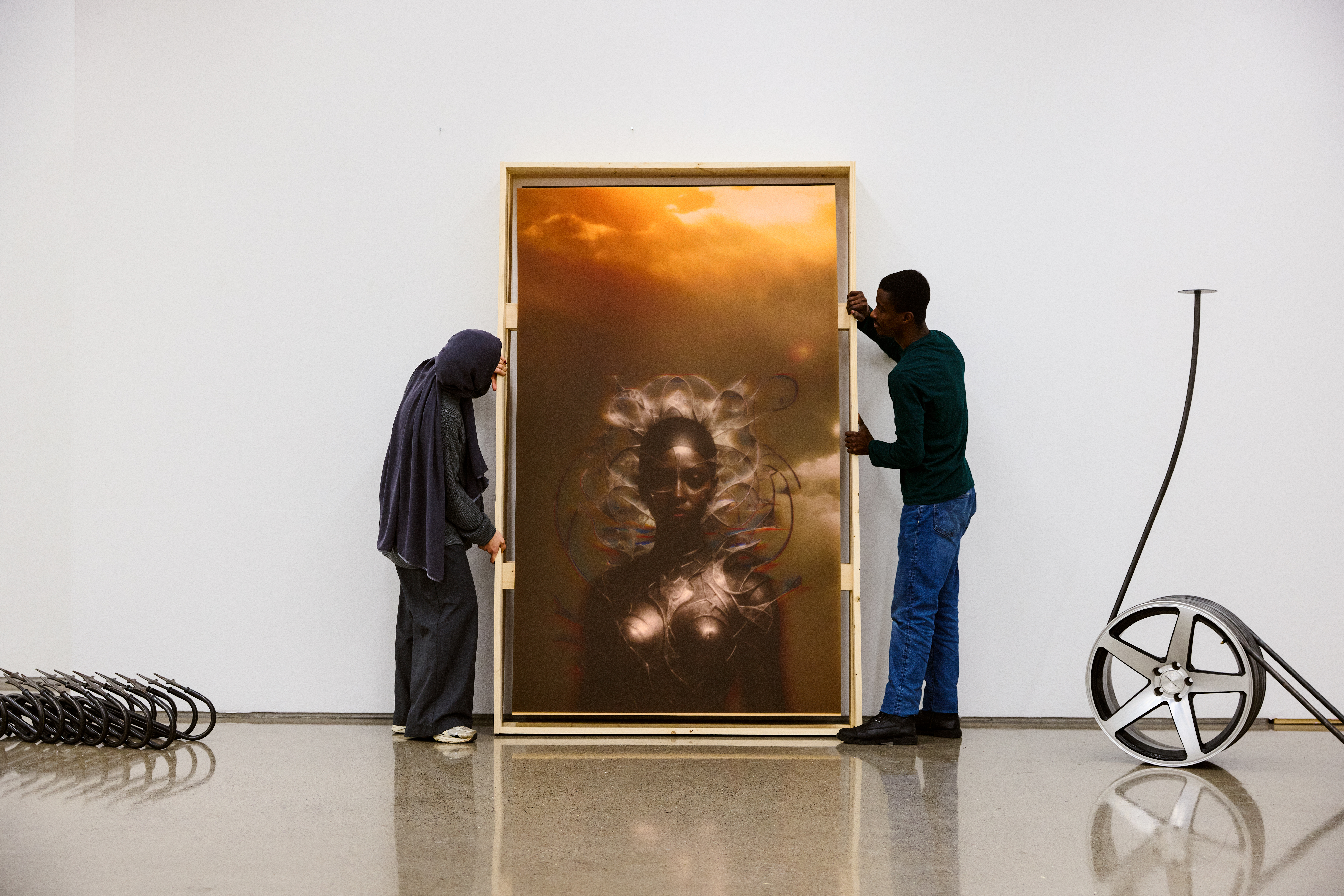 To personer plasserer et stort innrammet kunstverk som avbilder en mystisk figur mot en solnedgangshimmel. Bilhjul og metalldeler er synlige i nærheten.