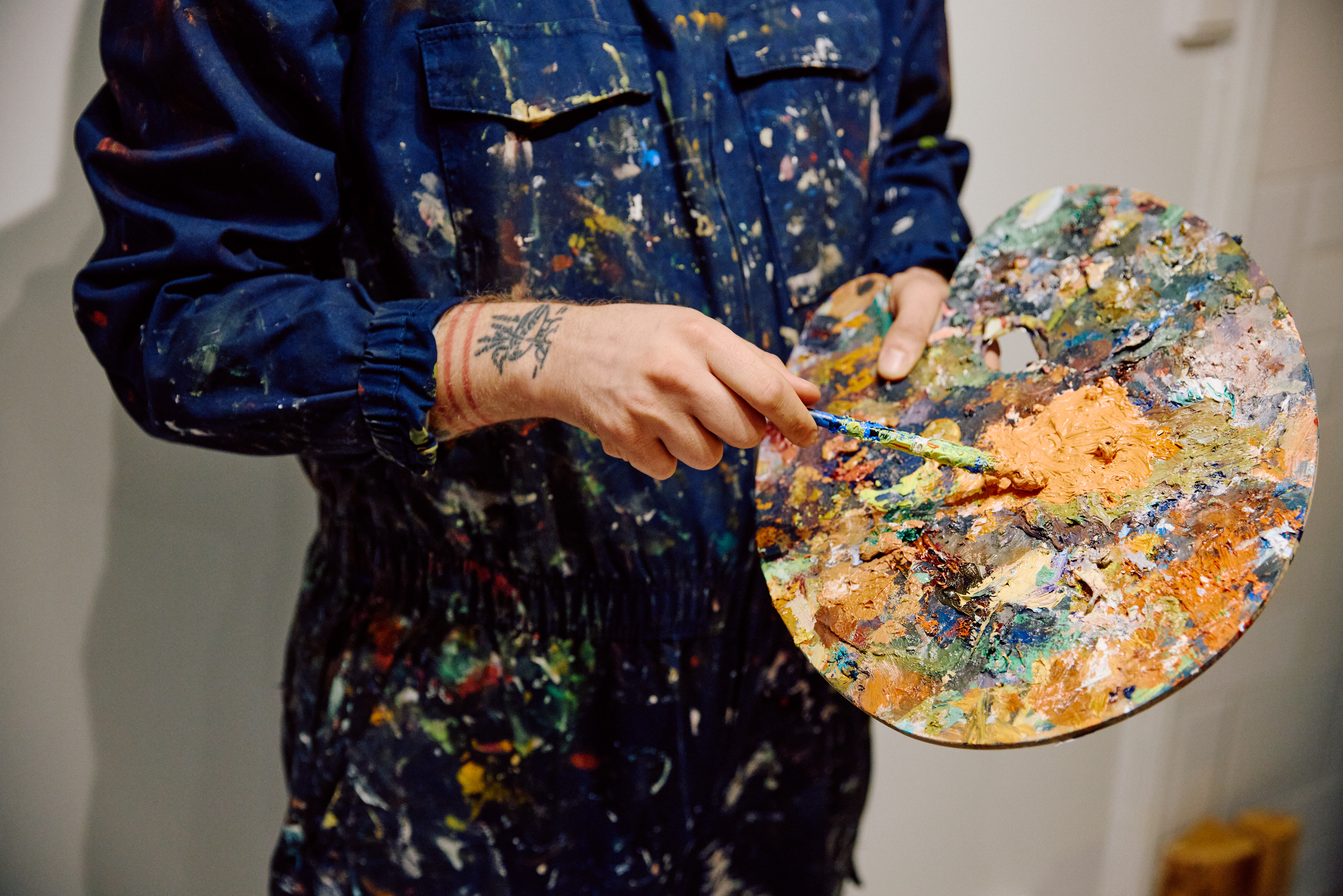 Kunstner i malingssprutet blå kjeledress som holder en fargerik palett, med tatoverte hender som blander maling til kunstverket sitt.