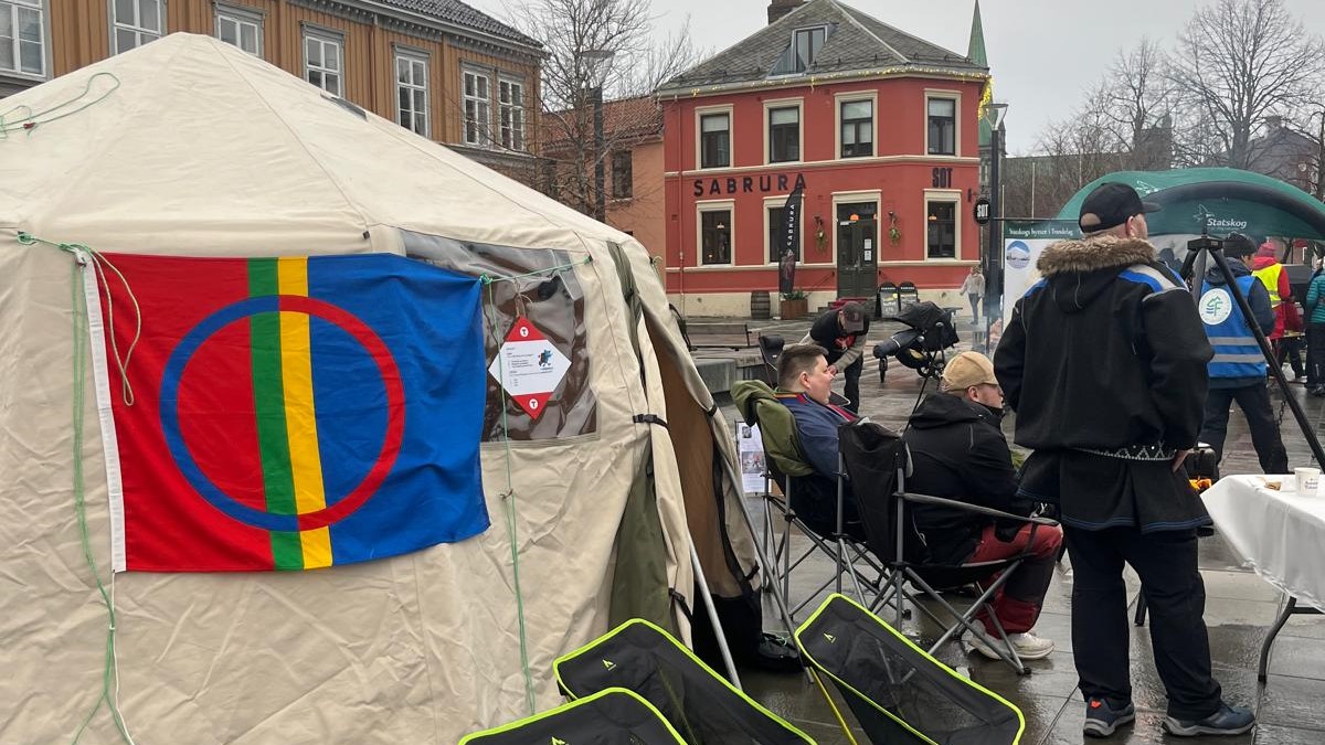 En offentlig samling eller demonstrasjon i en by, med en stor beige telt i forgrunnen og det samiske flagget festet til teltet. Flere personer sitter i klappstoler eller står og snakker sammen. I bakgrunnen sees bygninger, blant annet med skiltet "SABRURA", og en grønn oppblåsbar struktur med tekst. Været er overskyet og vått, og settingen tyder på en kulturell eller politisk markering, sannsynligvis knyttet til samiske rettigheter eller miljøspørsmål.