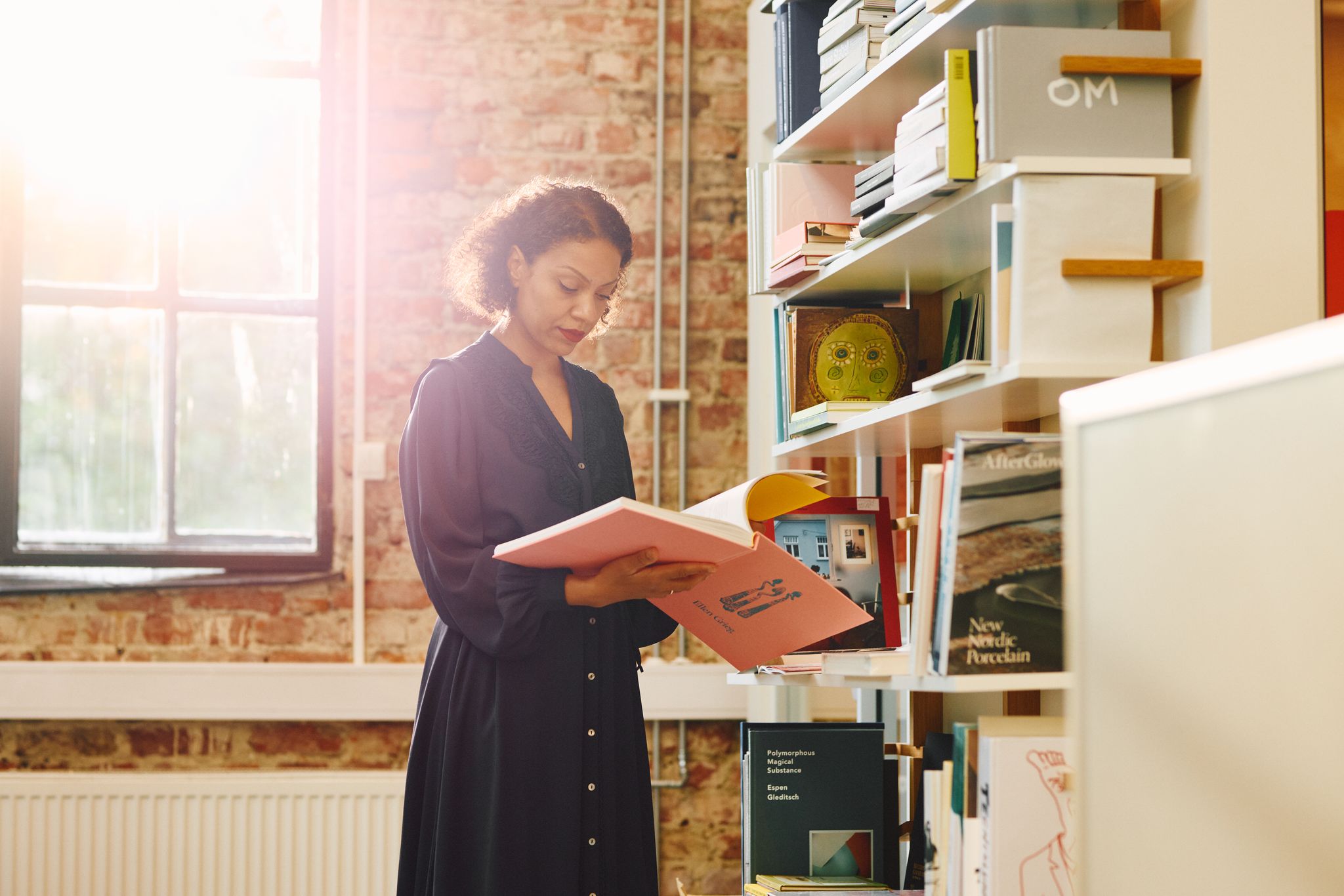 En kvinne står midt i bildet og blar i en rosa bok. Bak henne strømmer sollys inn gjennom et vindu, og til høyre står en bokhylle med flere bøker.