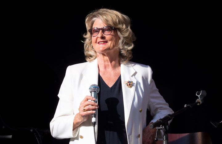 En person står på en mørk scene og holder en mikrofon i den ene hånden, med en ekstra mikrofon festet foran seg. Vedkommende har lyst, stylet hår, solbriller og en hvit blazer med dekorativ nål, samt mørkt antrekk under. Lyssettingen fremhever ansiktet og klærne, og skaper et tydelig fokus mot personen. Bildet formidler et øyeblikk av formidling eller opptreden i en profesjonell og stemningsfull setting.