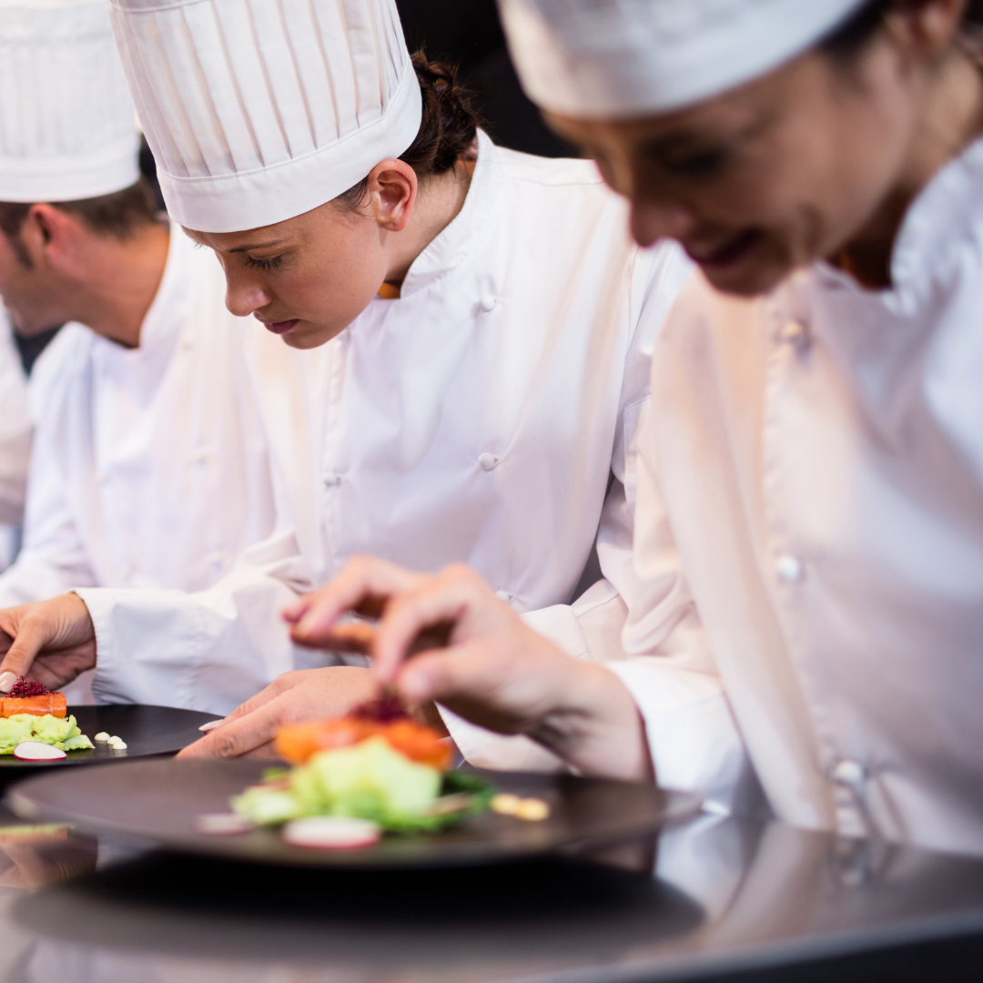 A medium close up of three young chefs stood in a row plating food onto black dishes.
