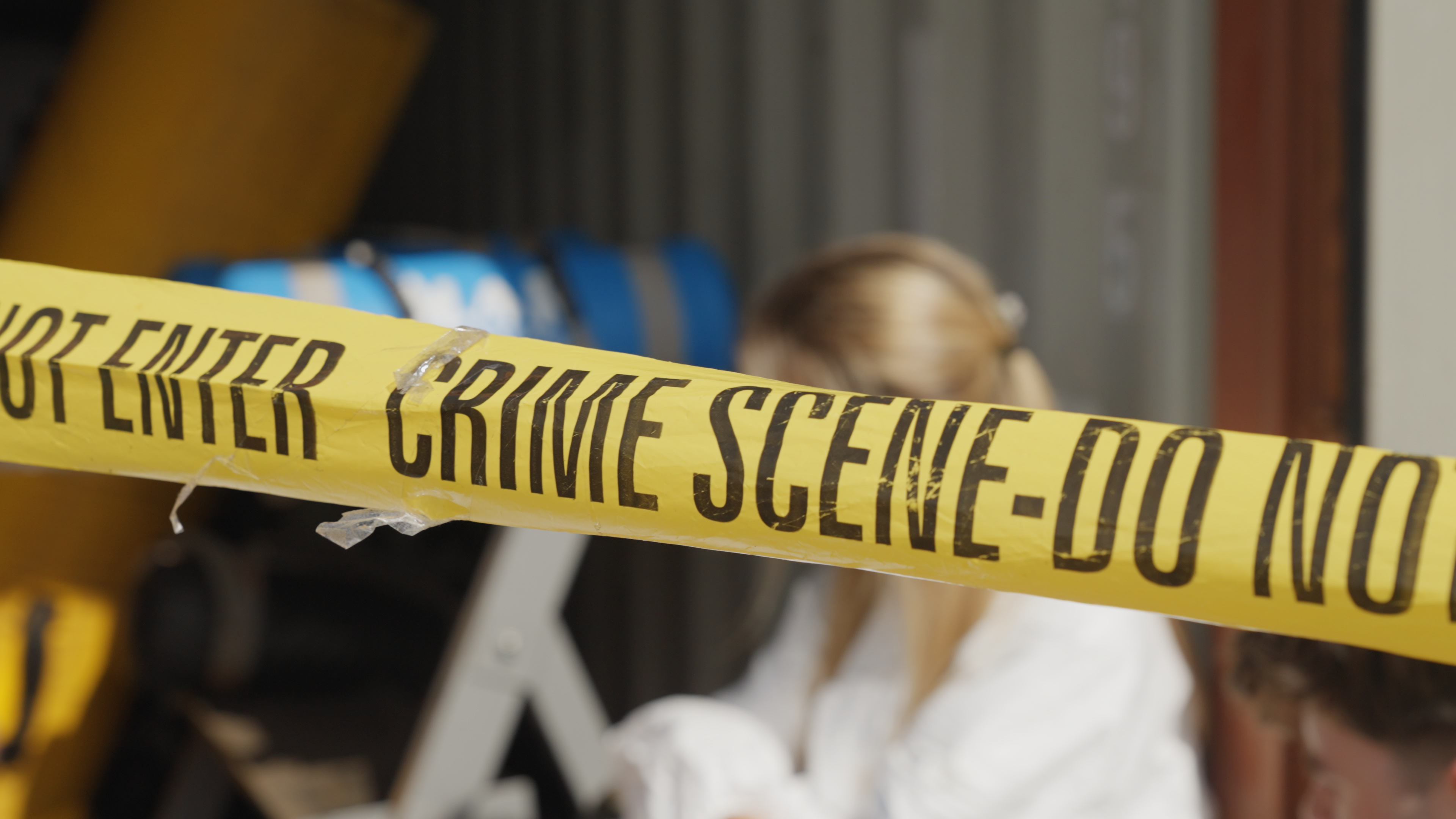 A medium close-up of yellow "Crime scene - do not cross" tape. A blurred individual is crouched down behind the tape looking off to the left.