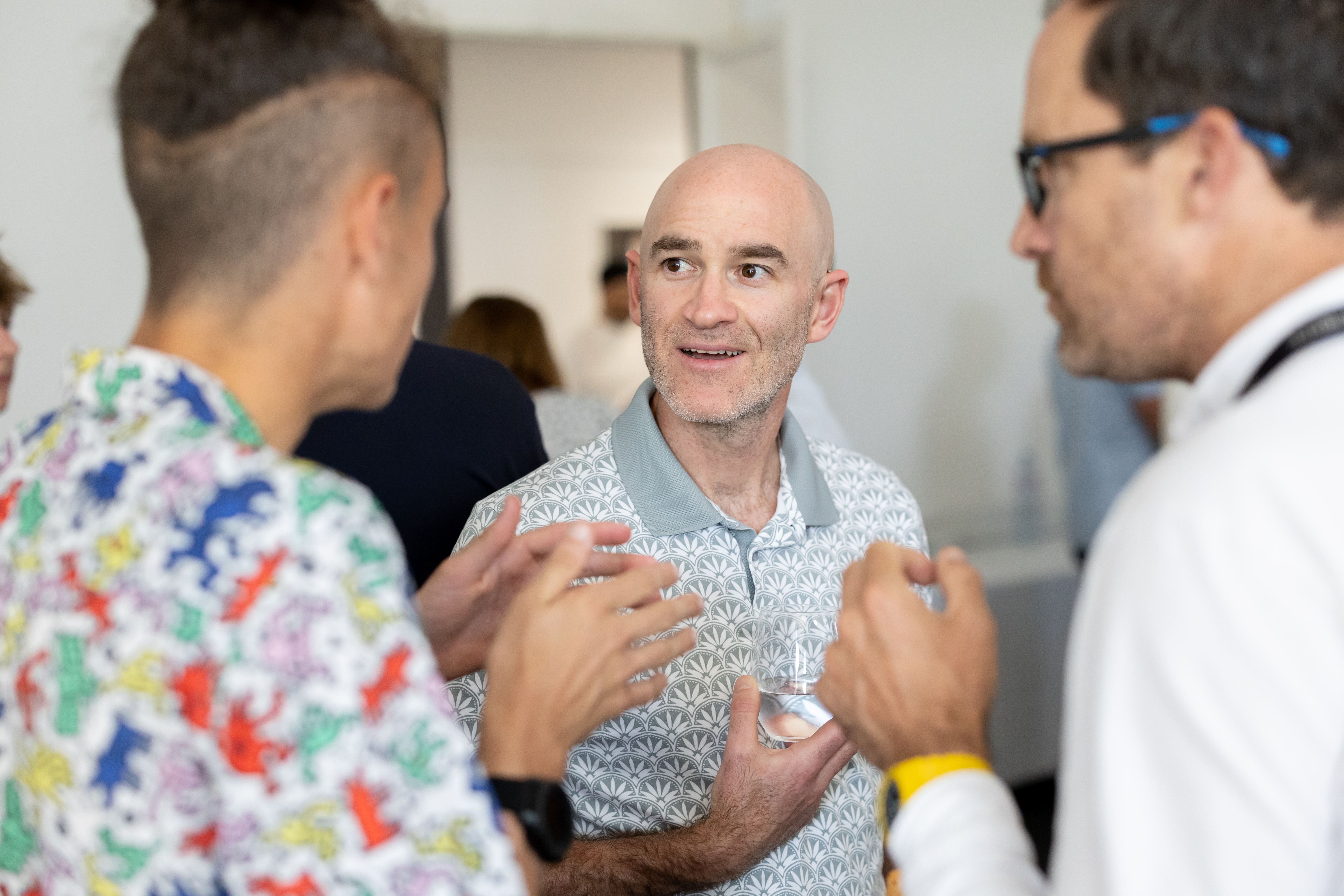 A bald man in a patterned polo shirt holding a glass, engaged in a conversation between two other men at an indoor event.