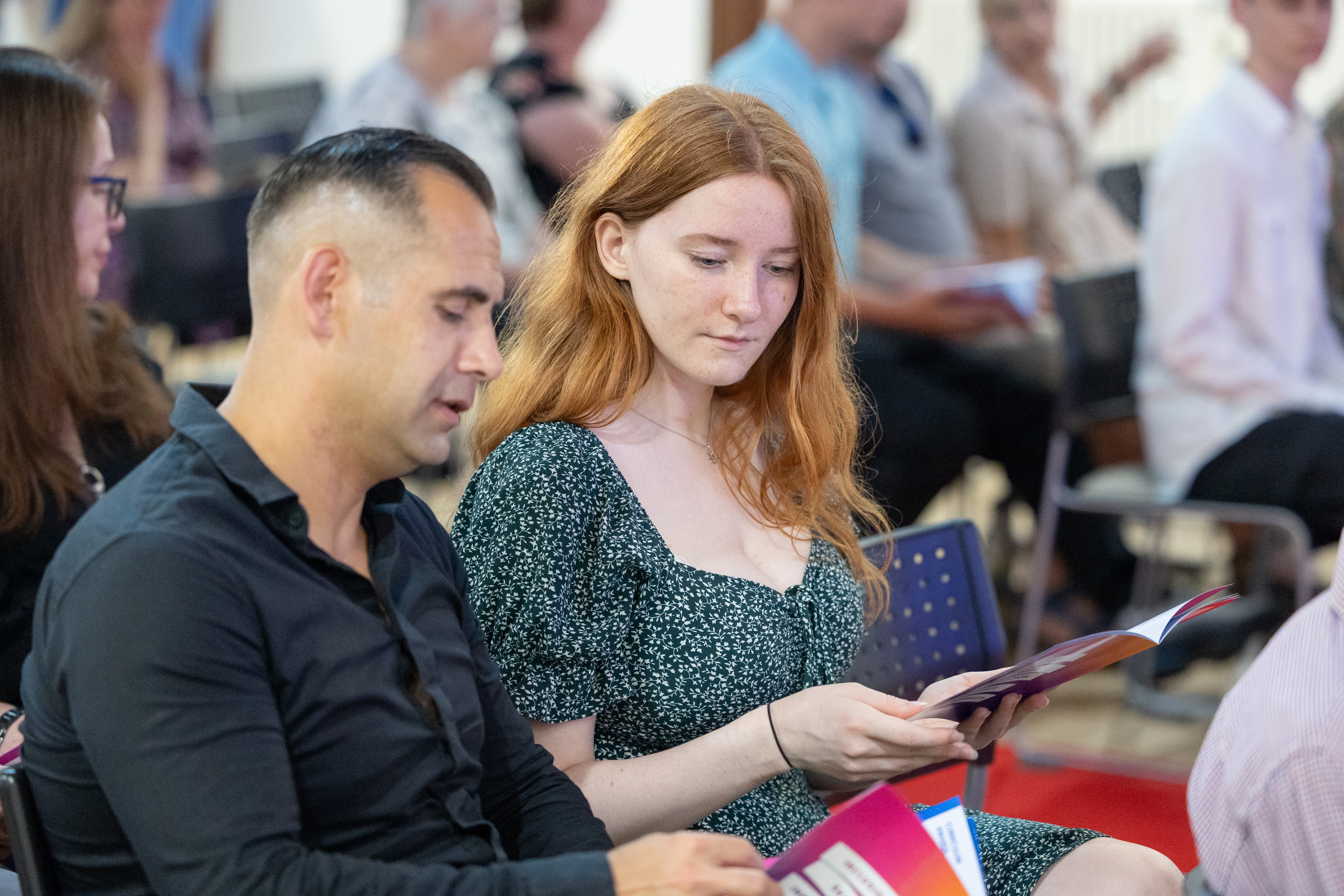 Two people who appear to be a father and daughter sitting in an audience, looking at event programmes during a ceremony