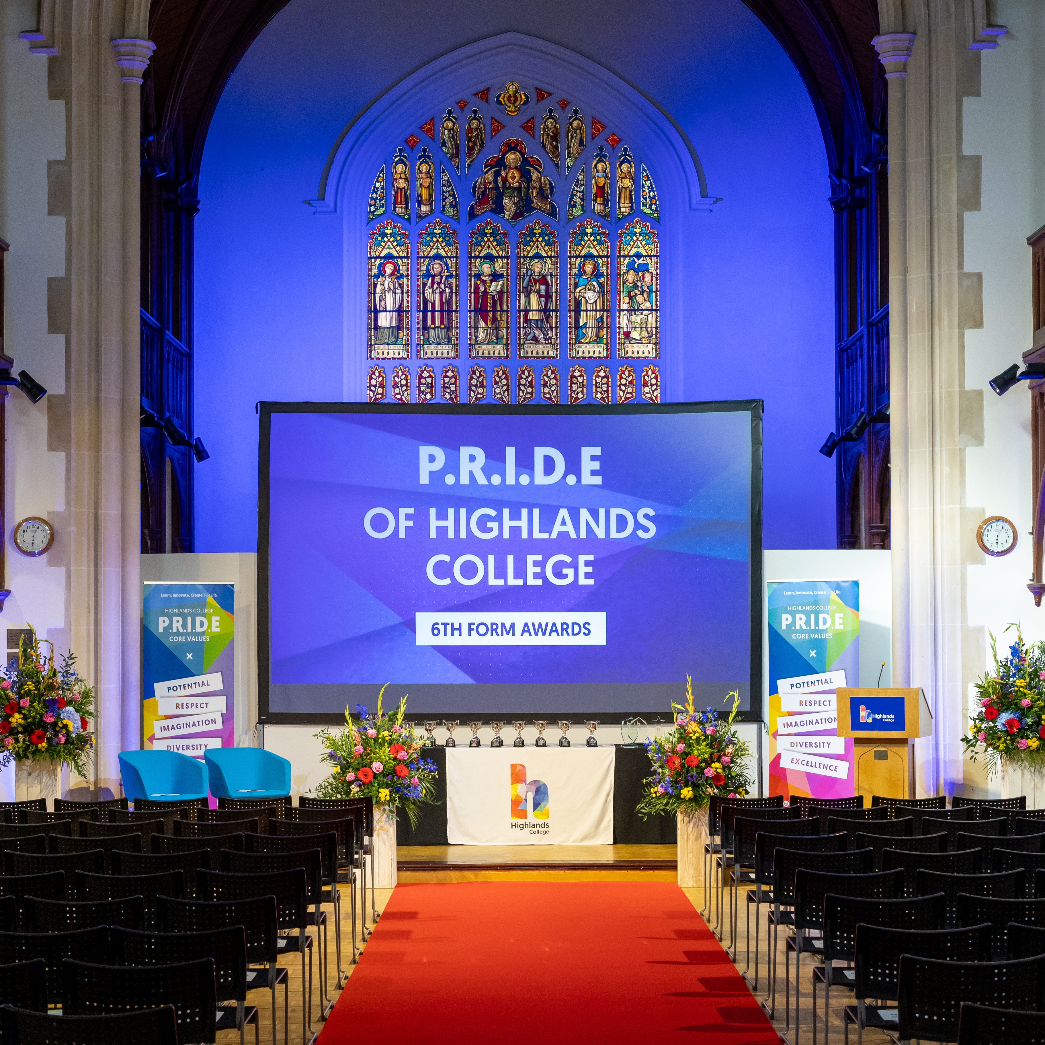 The Highlands College great hall set up with blue lighting on the stain-glass window and arch of the building, a red carpet leading to the stage with black seating facing the front on either side. The screen on stage reads PRIDE of Highlands College. Sixth Form Awards.