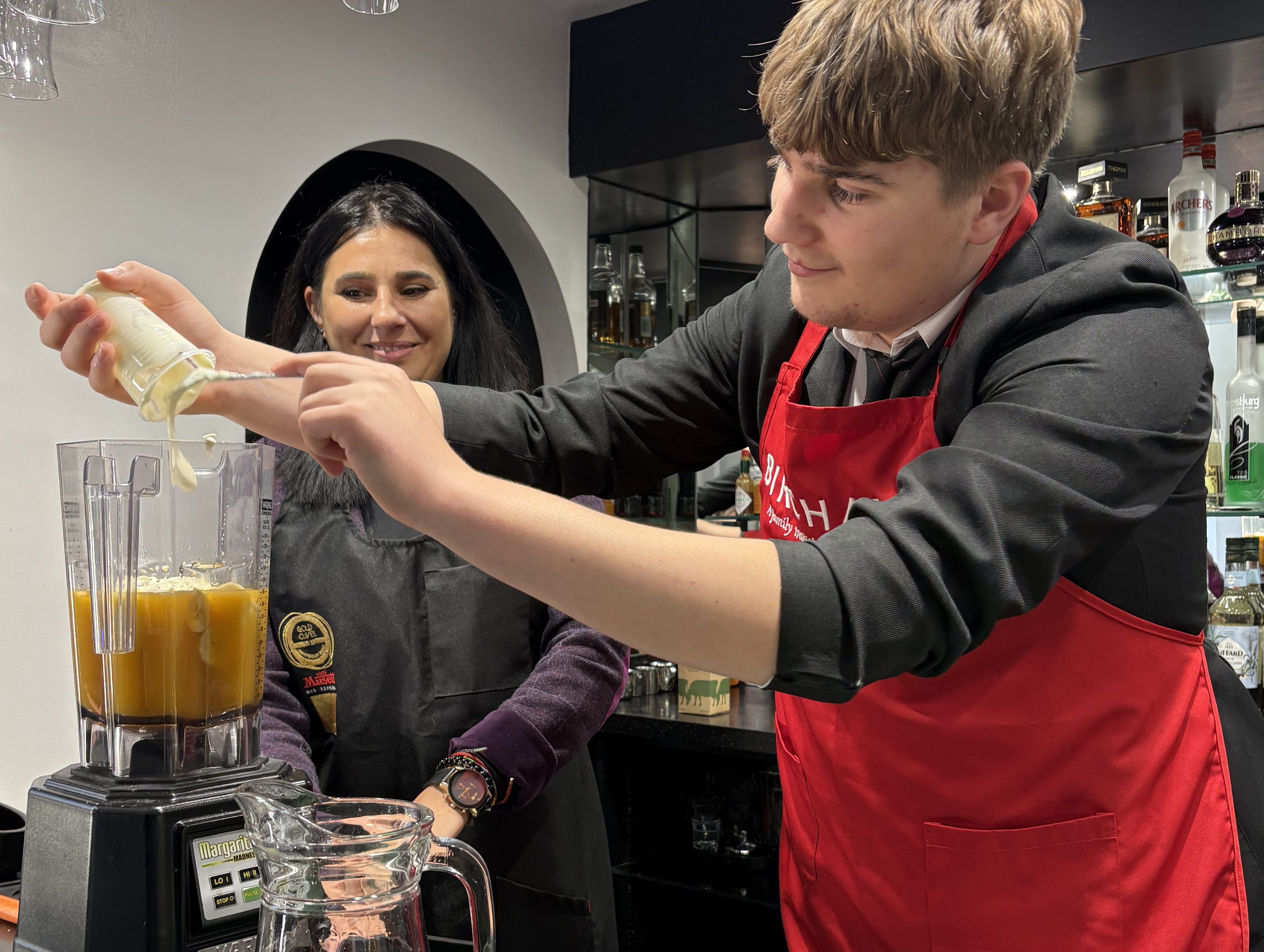 A male year 11 student behind the bar in our academy restaurant pouring yoghurt into a blender to make a smoothie. He's wearing a red apron and a black blazer under it. He's being supervised by a smiling member of staff.