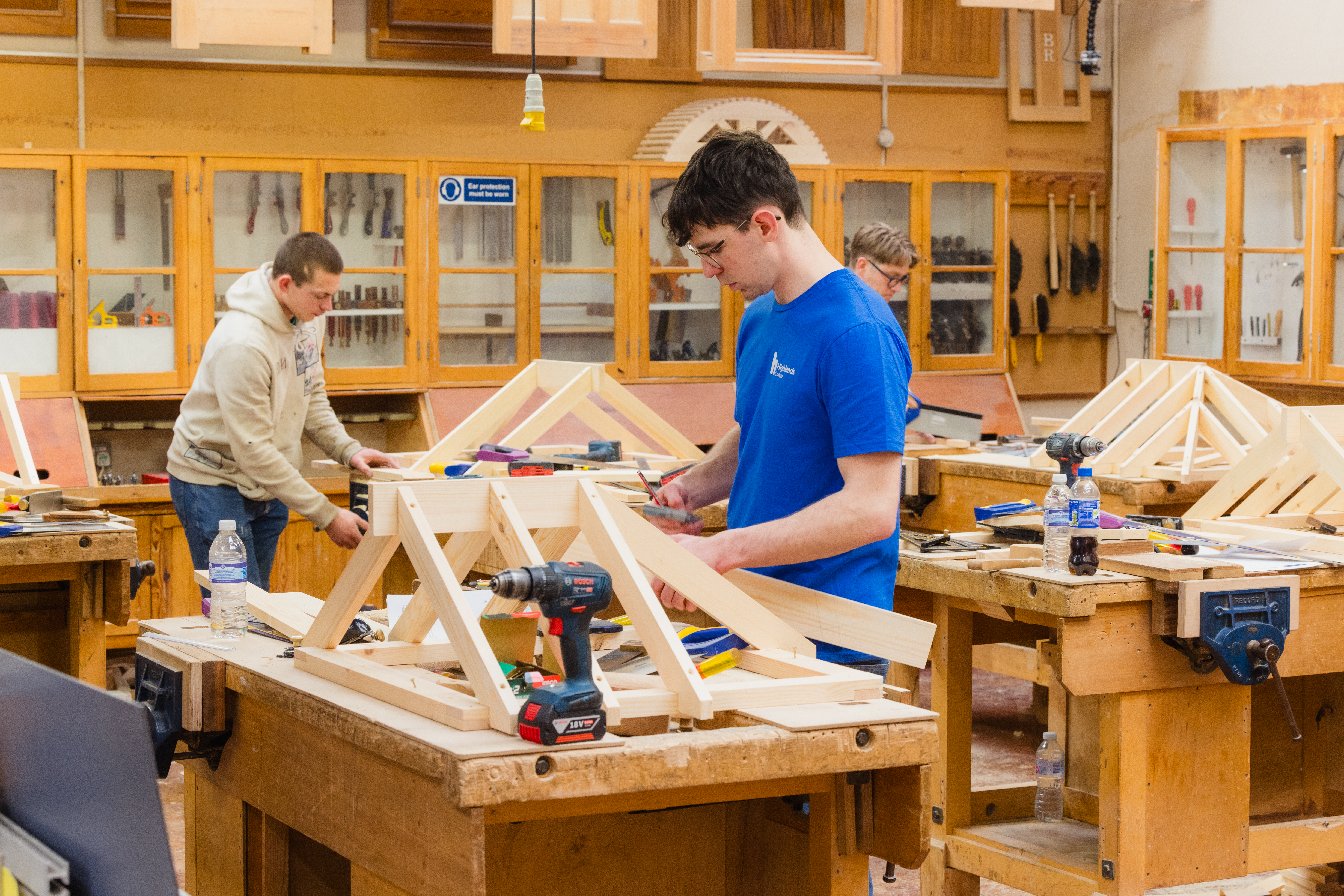 Three individuals working in a woodworking classroom, assembling wooden frames at benches surrounded by tools and materials.