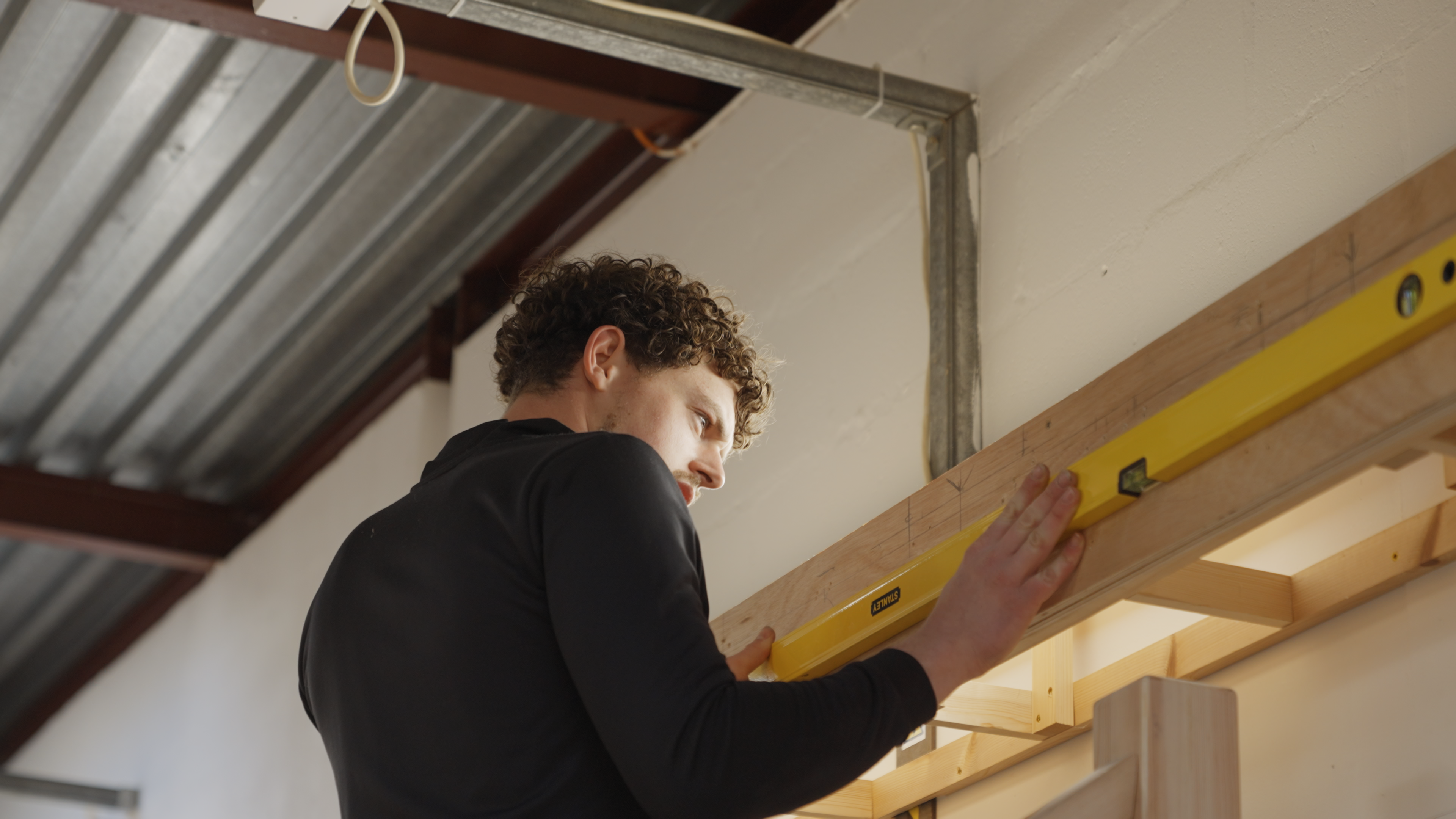 A medium shot of an individual holding a level against a wall-mounted wooden support. The wooden support is covered in pencil markings identifying measurements.