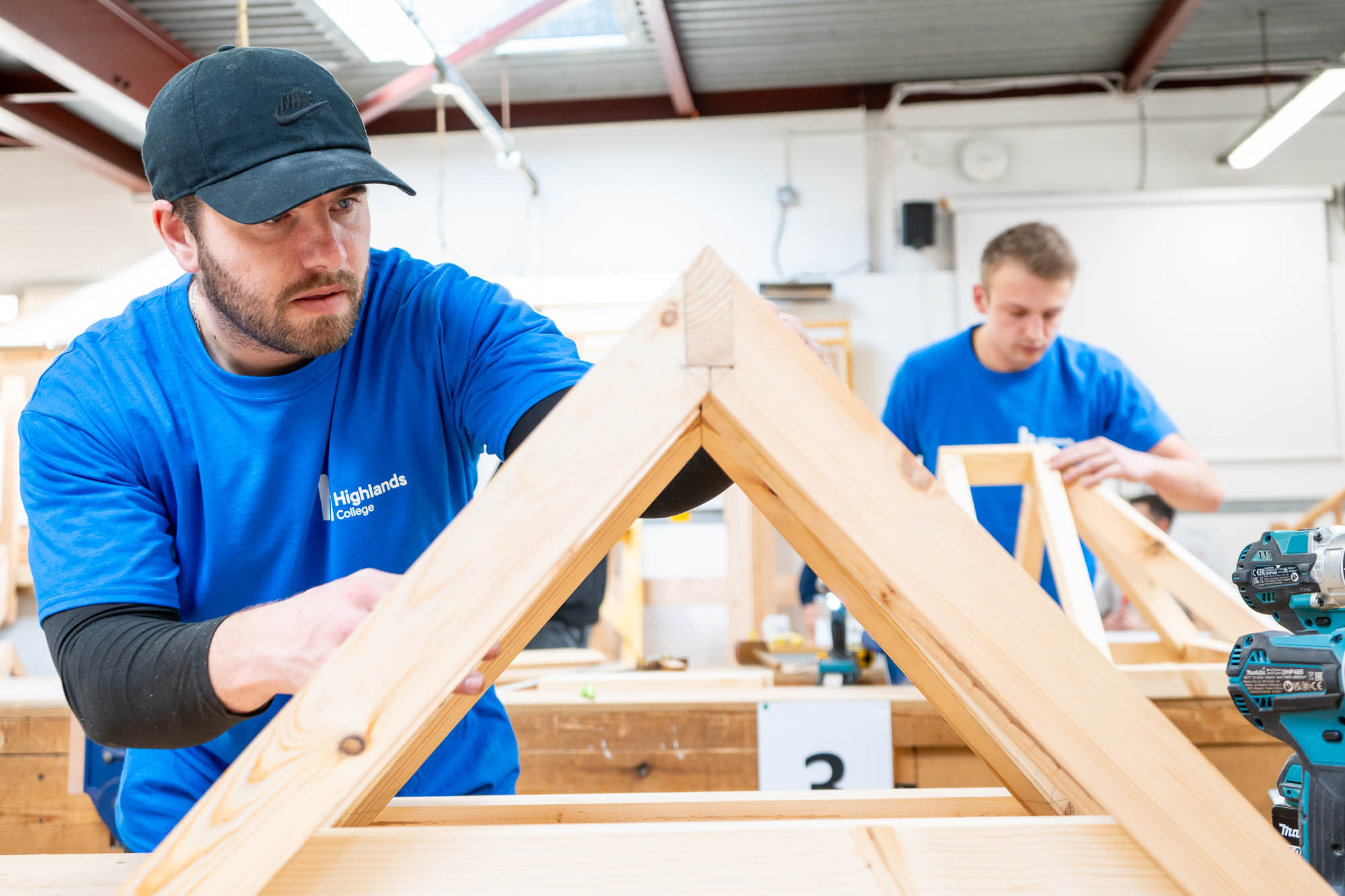 Two individuals in Highlands College shirts constructing wooden triangular frames in a workshop with tools and workbenches in the background.