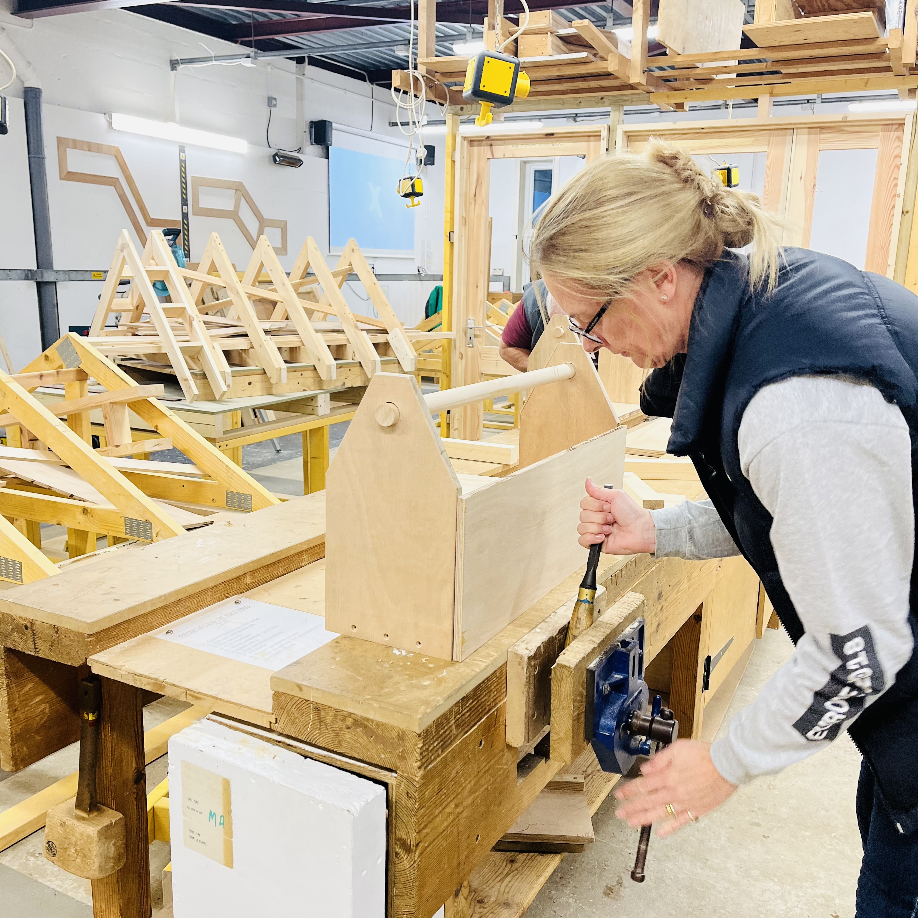 A photo of a woman in a wood workshop using a vice. She has a handmade wooden tool box in front of her on the workbench.