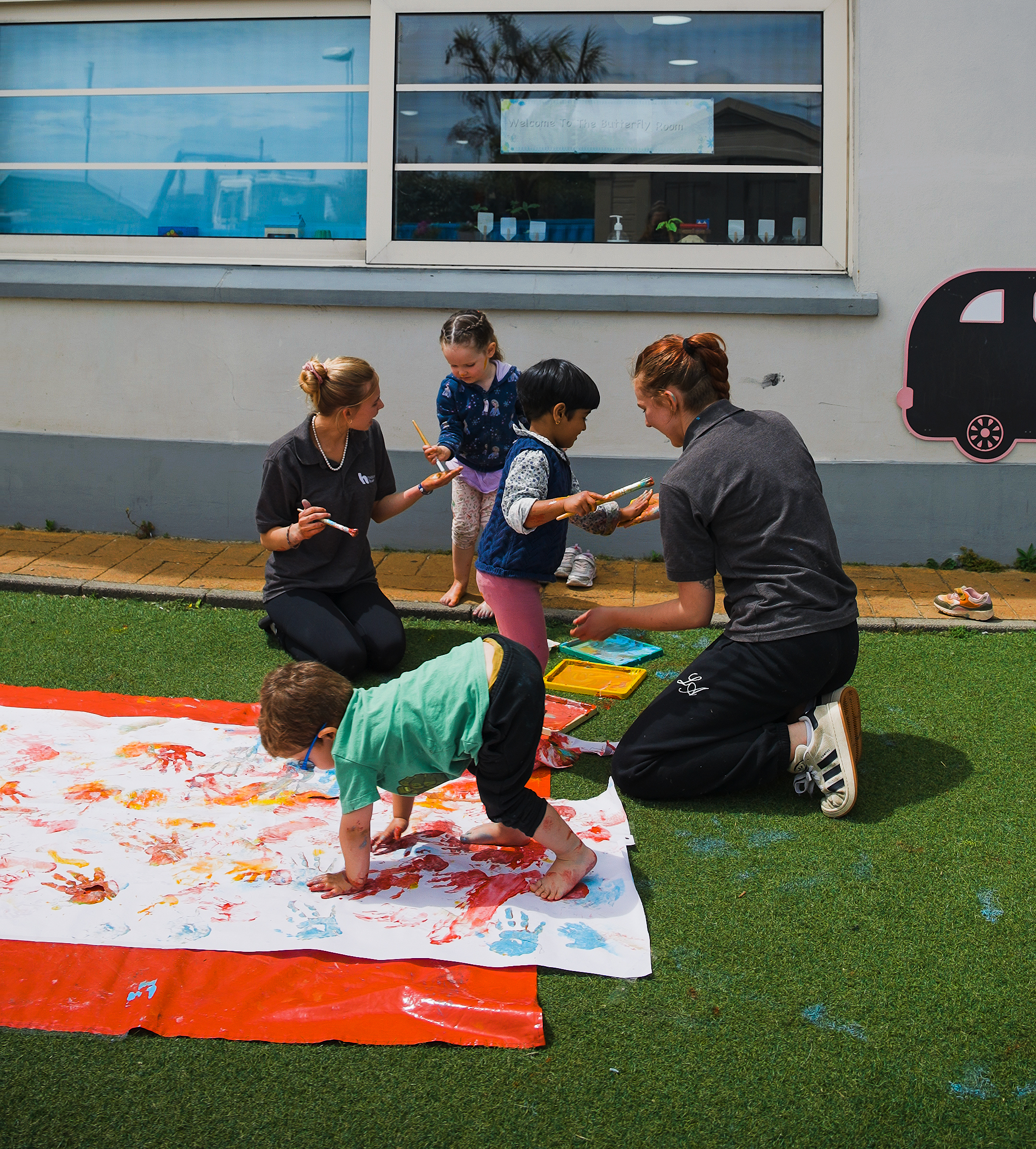 A wide photo of two students with three children playing with paint outside on a colourful matt.