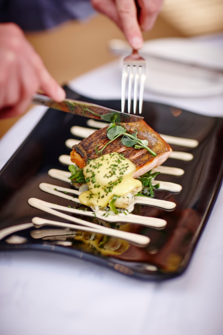 A close-up of a plated dish with seared salmon on greens, drizzled with creamy yellow sauce, being cut with a knife and fork on a black plate with white accents.