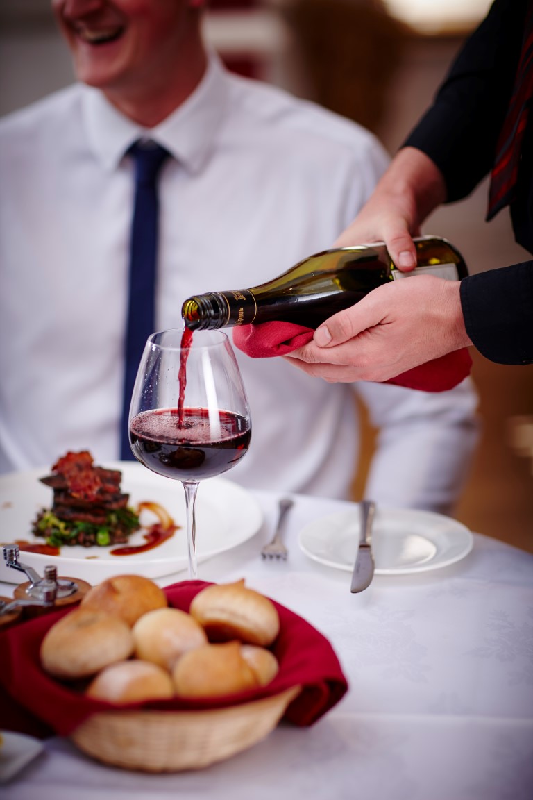 A medium close-up of a pair of hands pouring a glass of red wine for an individual seated at a dining table. There is a variety of food on the table.