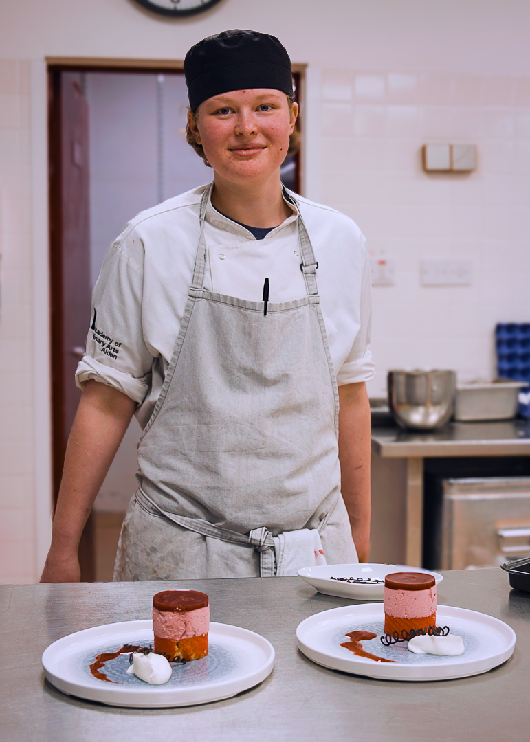 Apprenticeship chef in white uniform and grey apron standing behind a counter with three plated layered desserts, garnished with whipped cream and chocolate decorations in a kitchen setting.