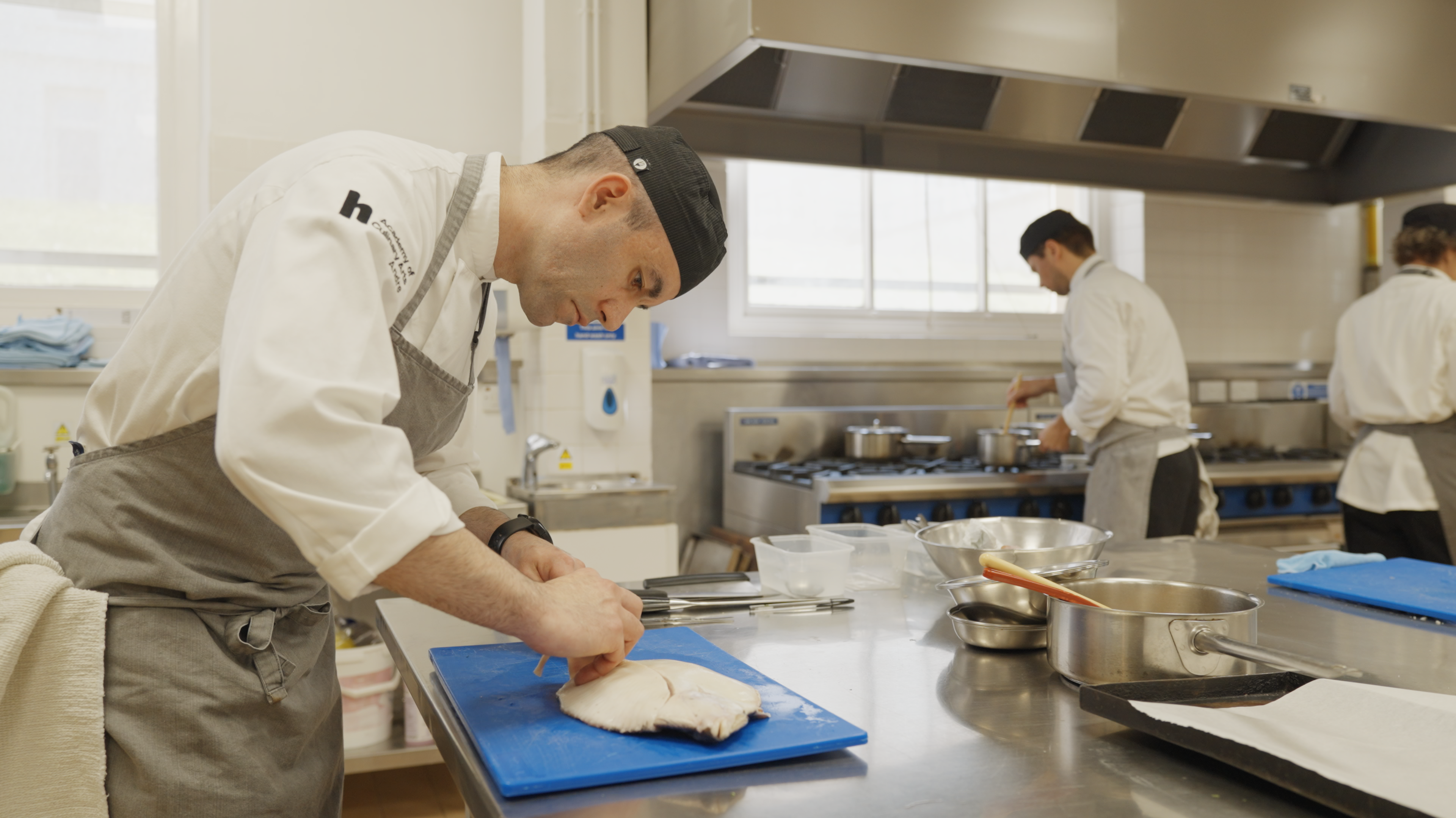 A medium shot of a kitchen with three chefs. The chef in the foreground is filleting a flat fish on a blue chopping board.