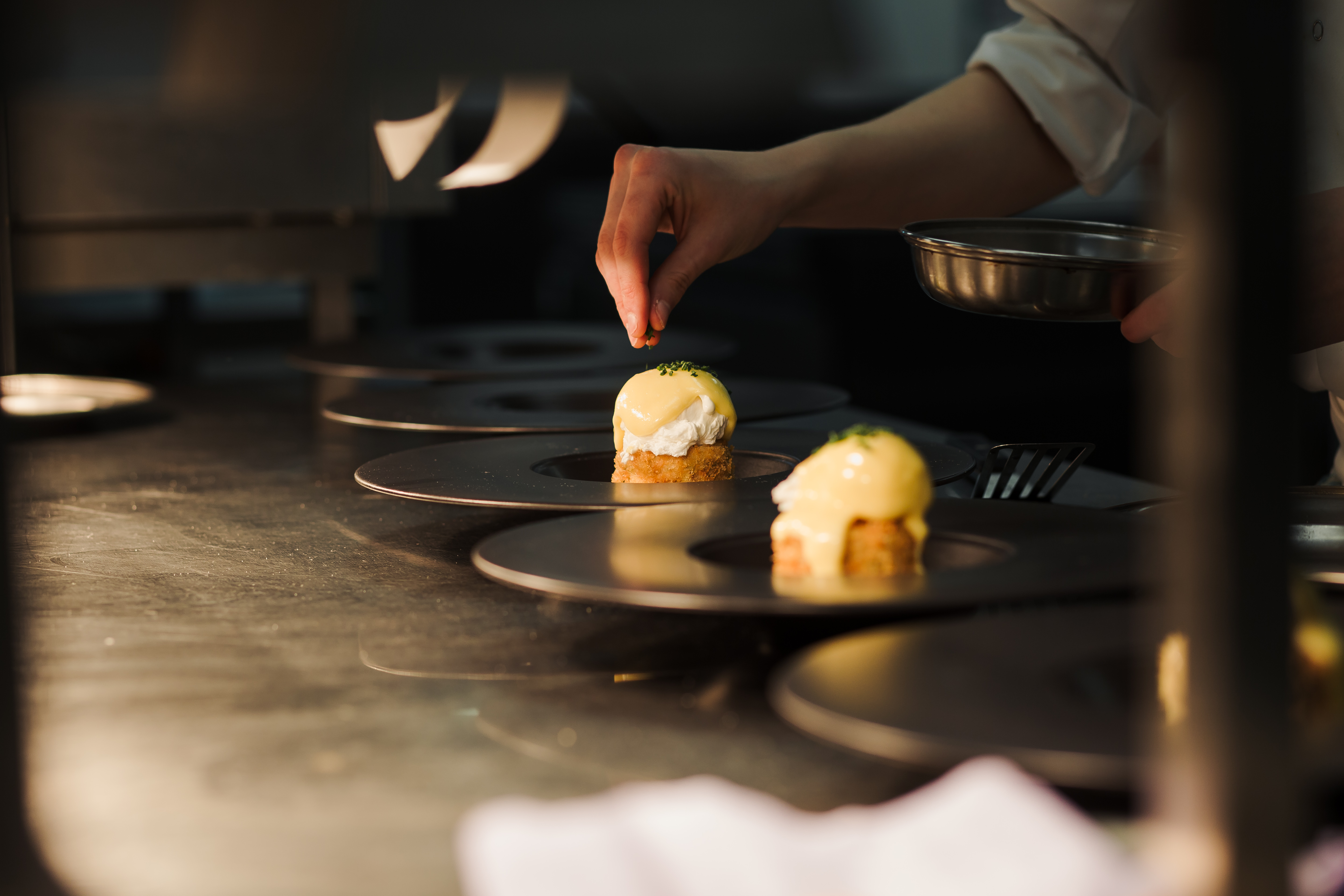 Chef plating dishes in a professional kitchen, arranged on plates lined up on a counter.
