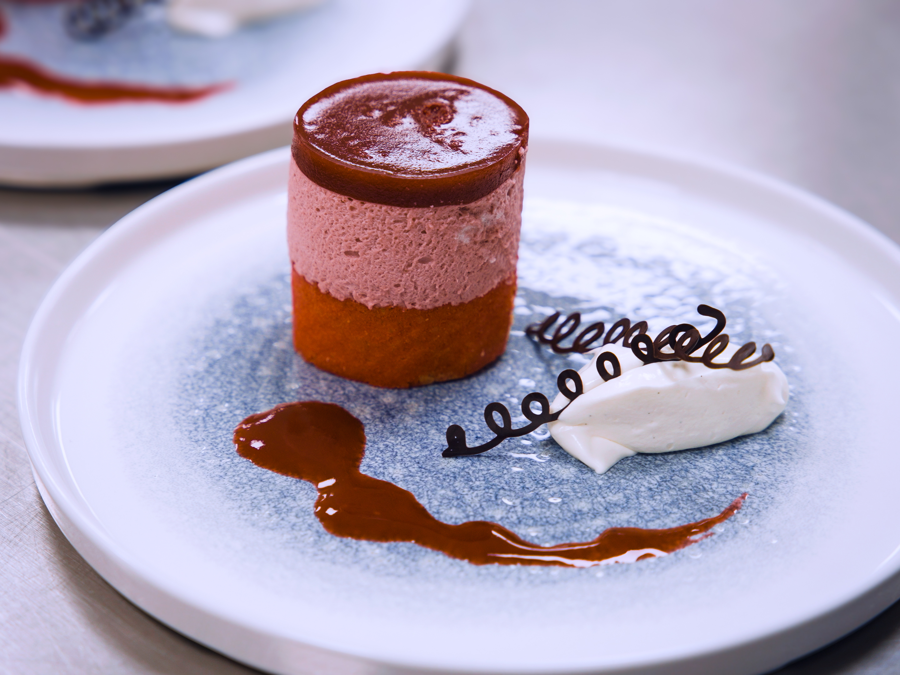 A close-up of a three layered dessert with vanilla ice cream on the side and decorative sauce plated on a round lipped blue and white plate.