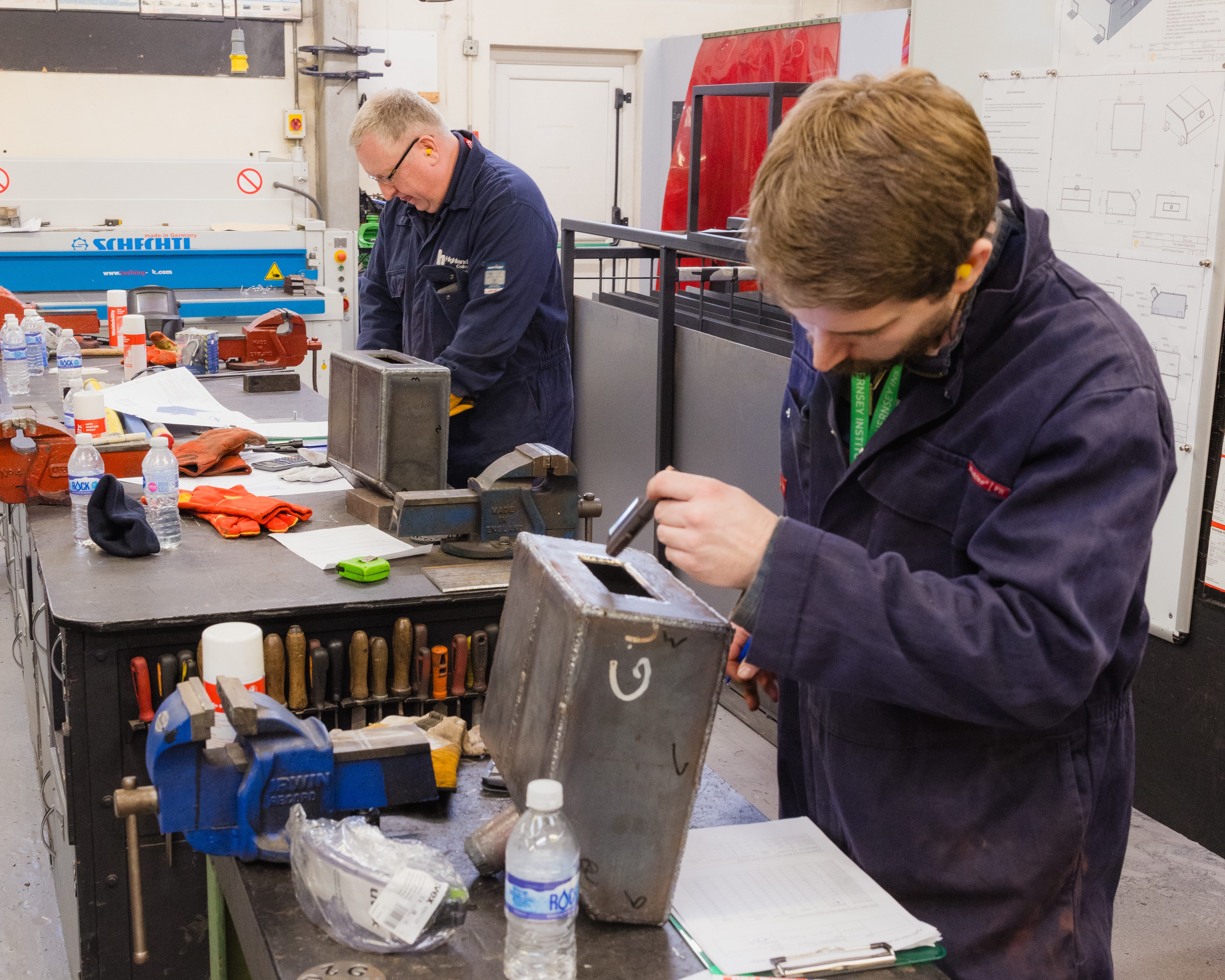 A medium shot of two individuals in protective clothing working in a technical workshop, handling metal boxes on a cluttered workbench with tools, gloves, and diagrams visible in the background.