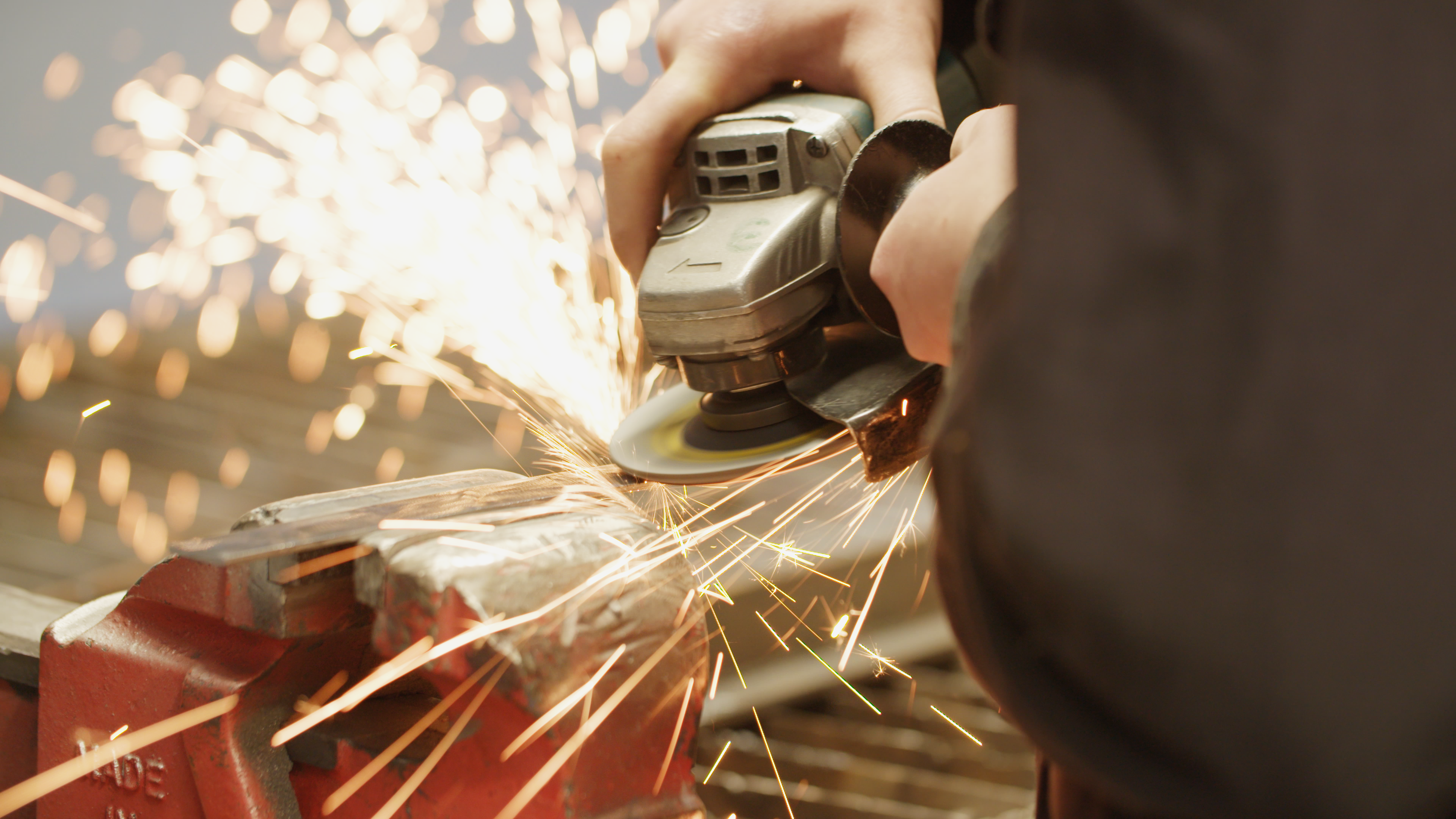 A close up of a pair of hands using an angle grinder on a piece of metal. There are sparks flying away from the hands.