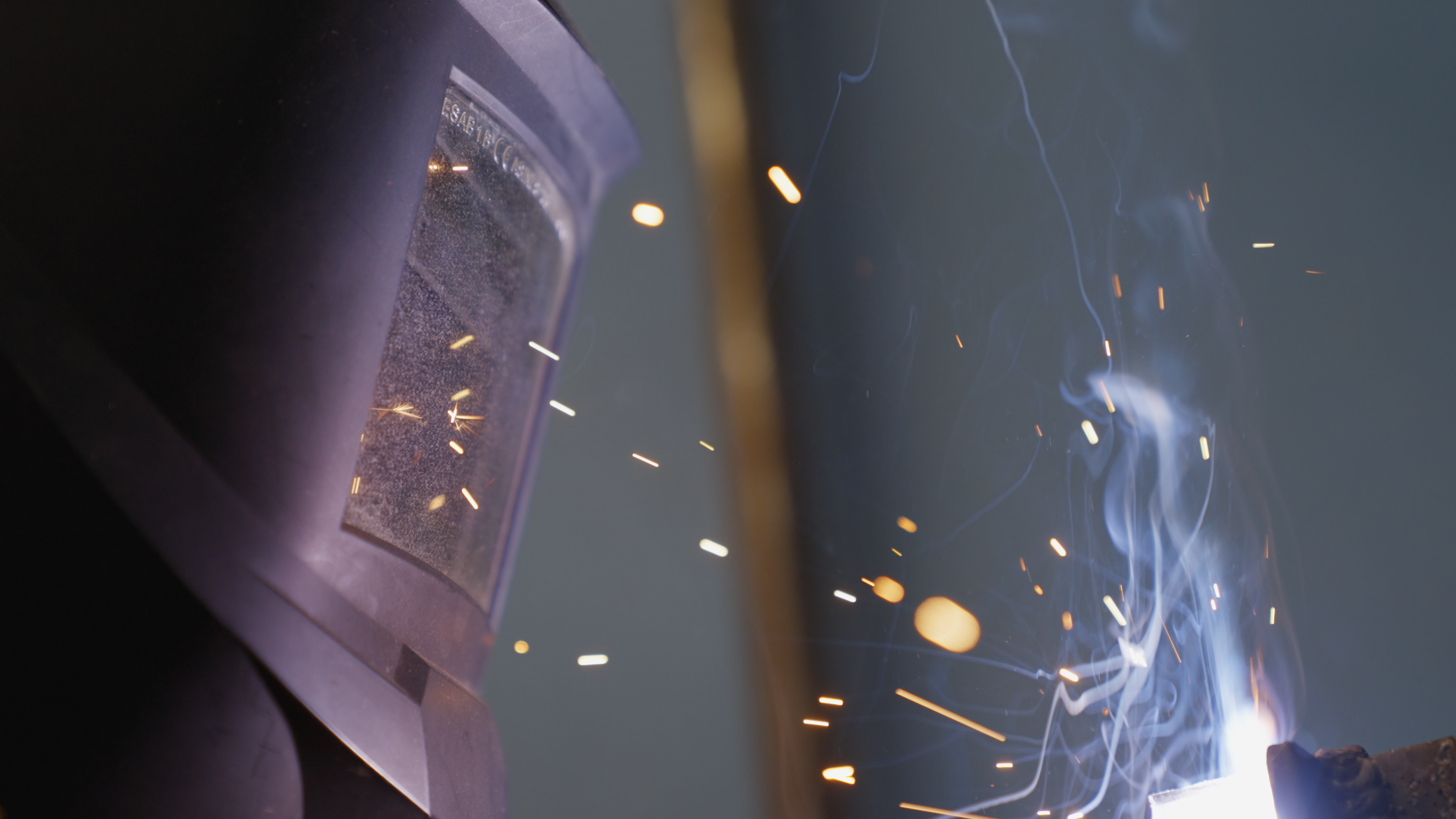 A close up of an individual wearing a welding mask. The individual is welding something out of frame, and sparks are flying towards the mask.