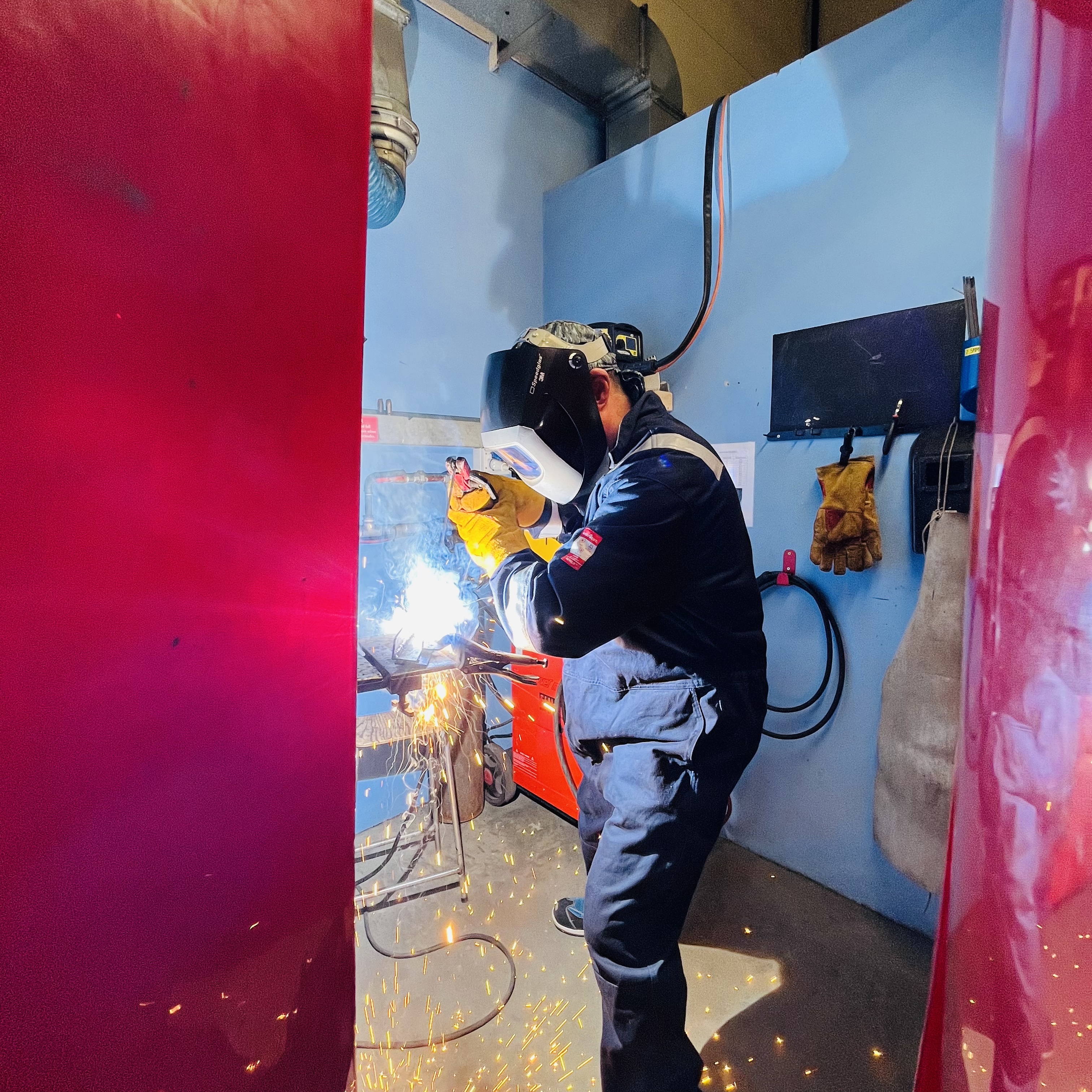 A medium shot of a person welding in a mask, with sparks flying. The individual is wearing blue overalls
