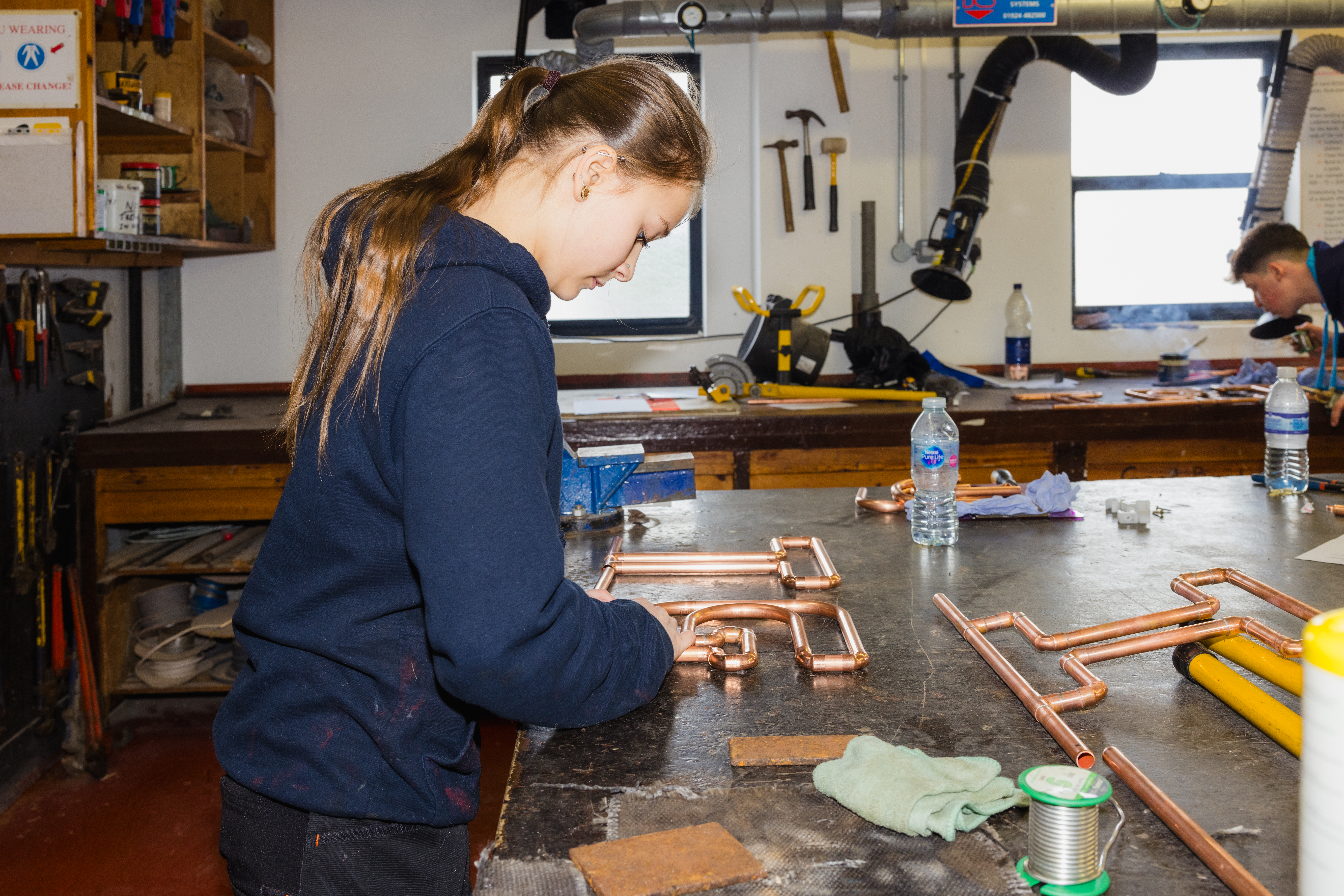 A medium shot of an individual in a workshop assembling copper pipes on a workbench, surrounded by tools, clamps, and soldering materials.