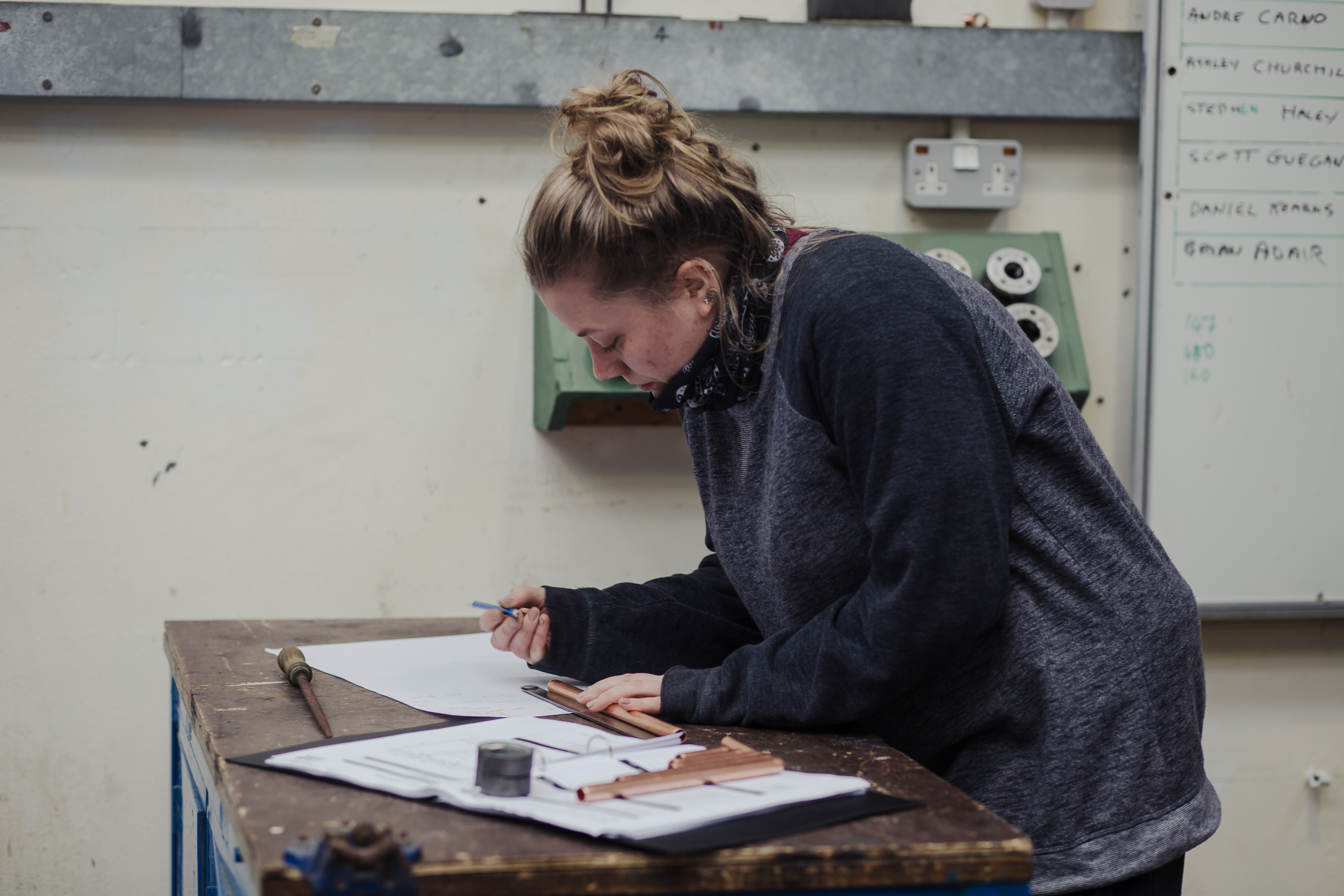 Person using drafting tools to work on a design at a workbench, with rulers, papers, and copper pipes visible; whiteboard in the background.