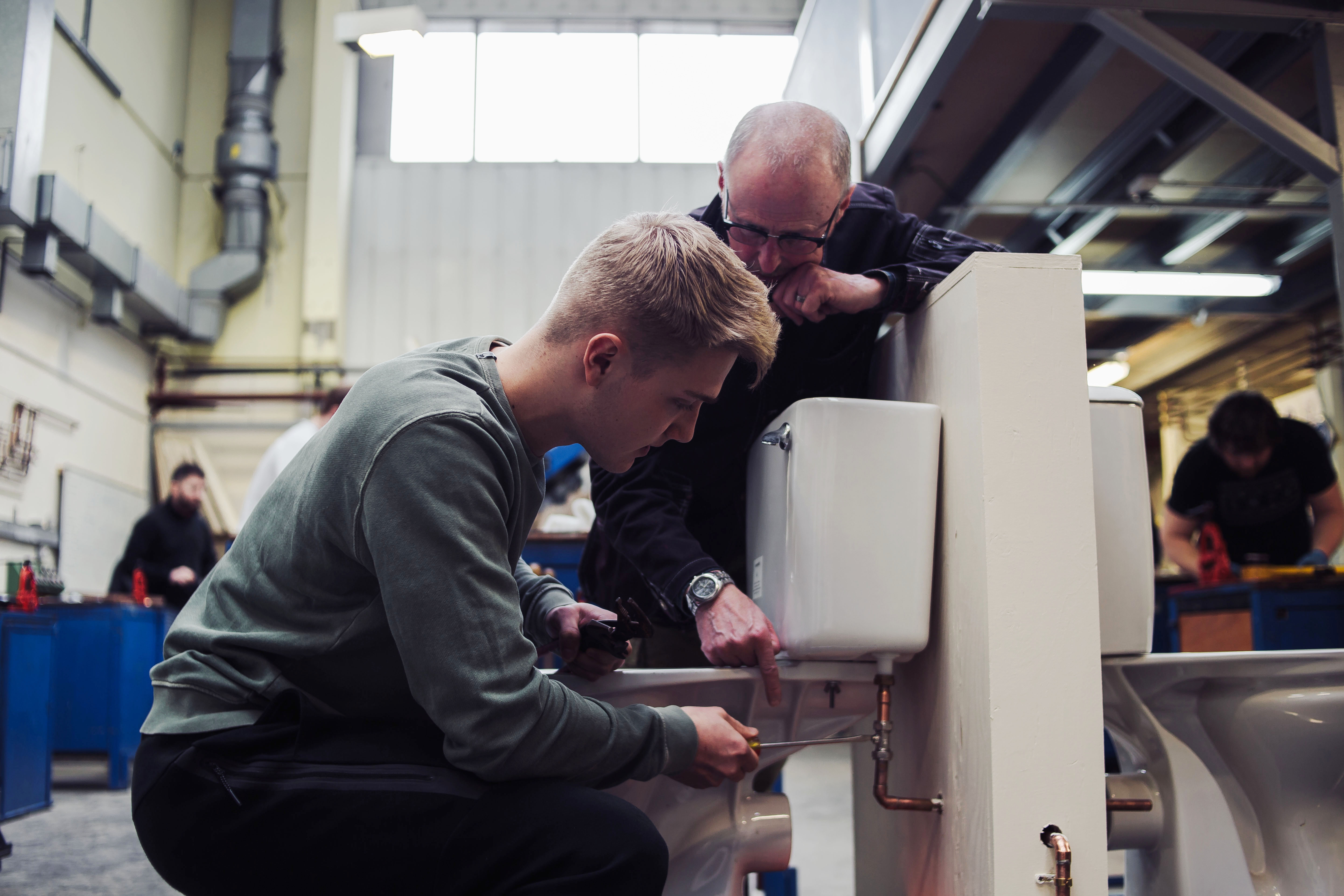 Two individuals working on a plumbing task in an industrial workshop; one kneeling with a tool, the other standing and assisting, with tools and others in the background.