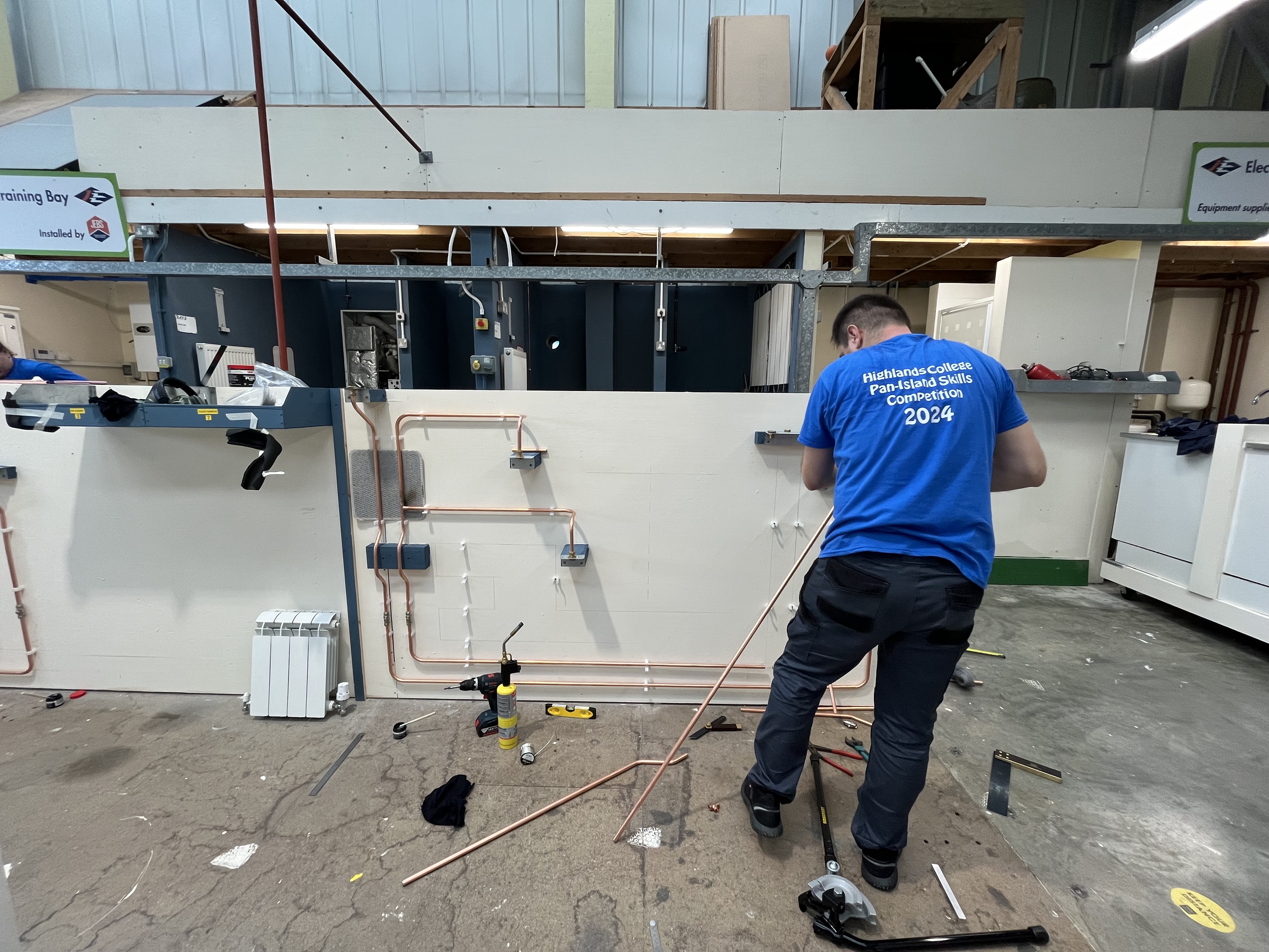 Person installing copper pipes on a white wall in a workshop, wearing a 'Plumbing College Future Skills Competition 2024' shirt, with tools and materials on the floor.