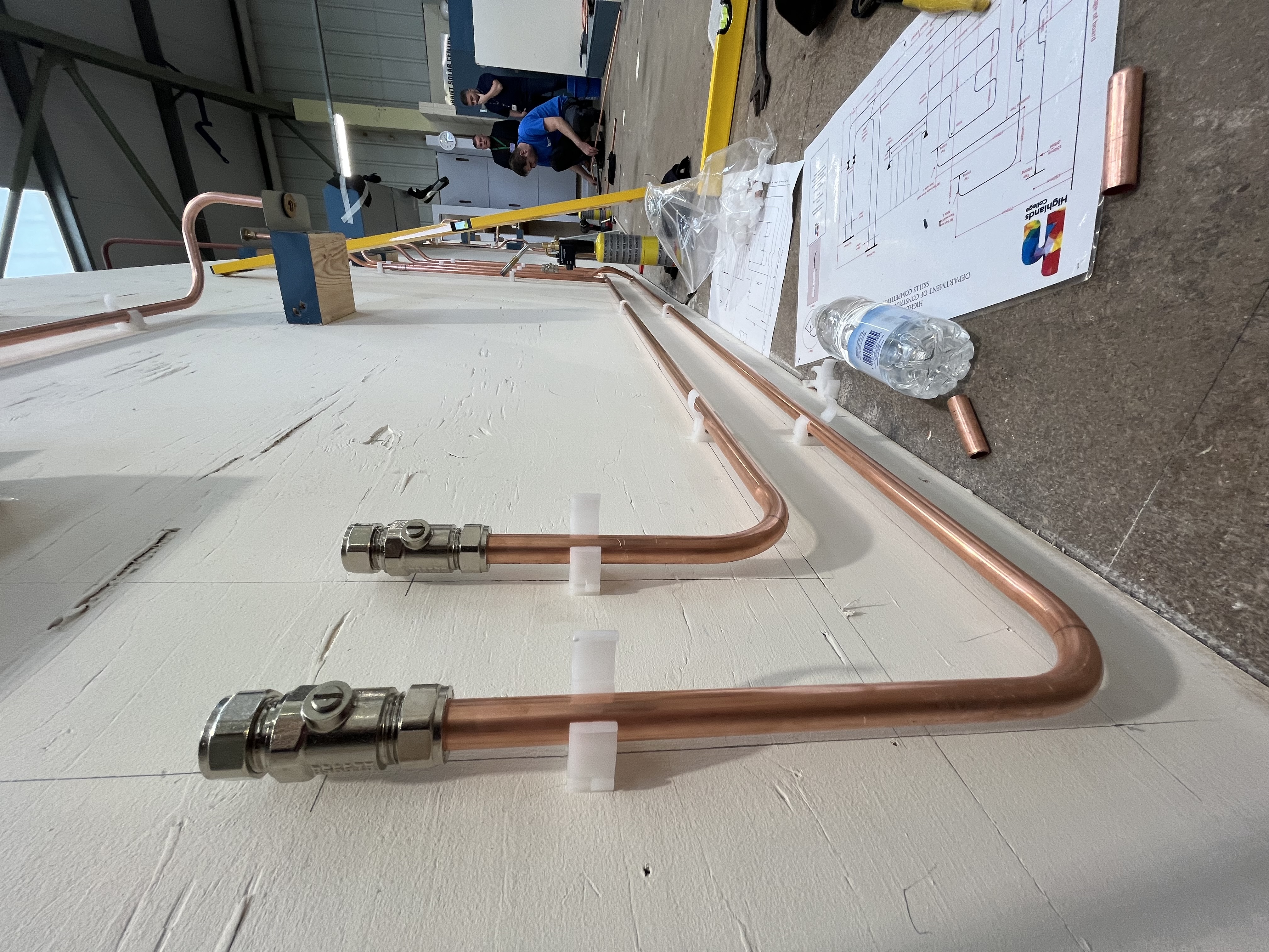 Copper pipes mounted on a white wall in a workshop, with fittings, brackets, and instructional papers on the floor, people and tools visible in the background.