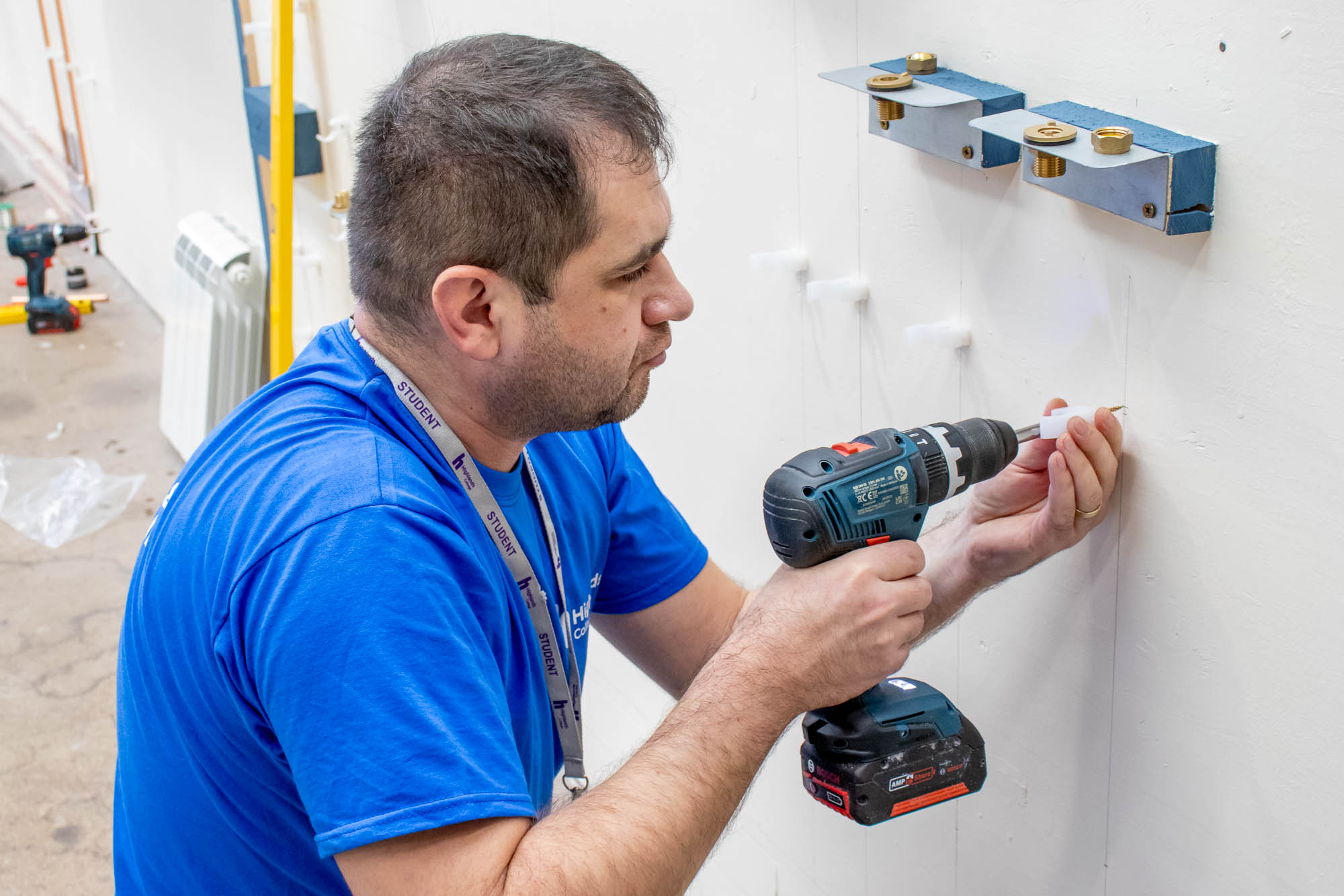 Person using a cordless drill on a white wall beneath mounted metal brackets, with tools and materials scattered on a concrete floor.