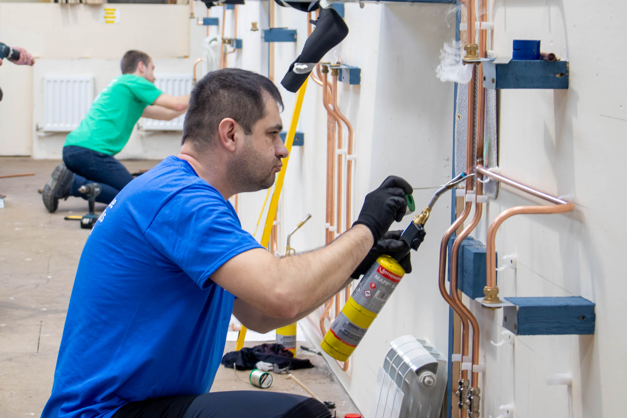 A medium wide shot of two individuals working on a plumbing system in a workshop; one soldering copper pipes with a blowtorch, the other kneeling with tools nearby.