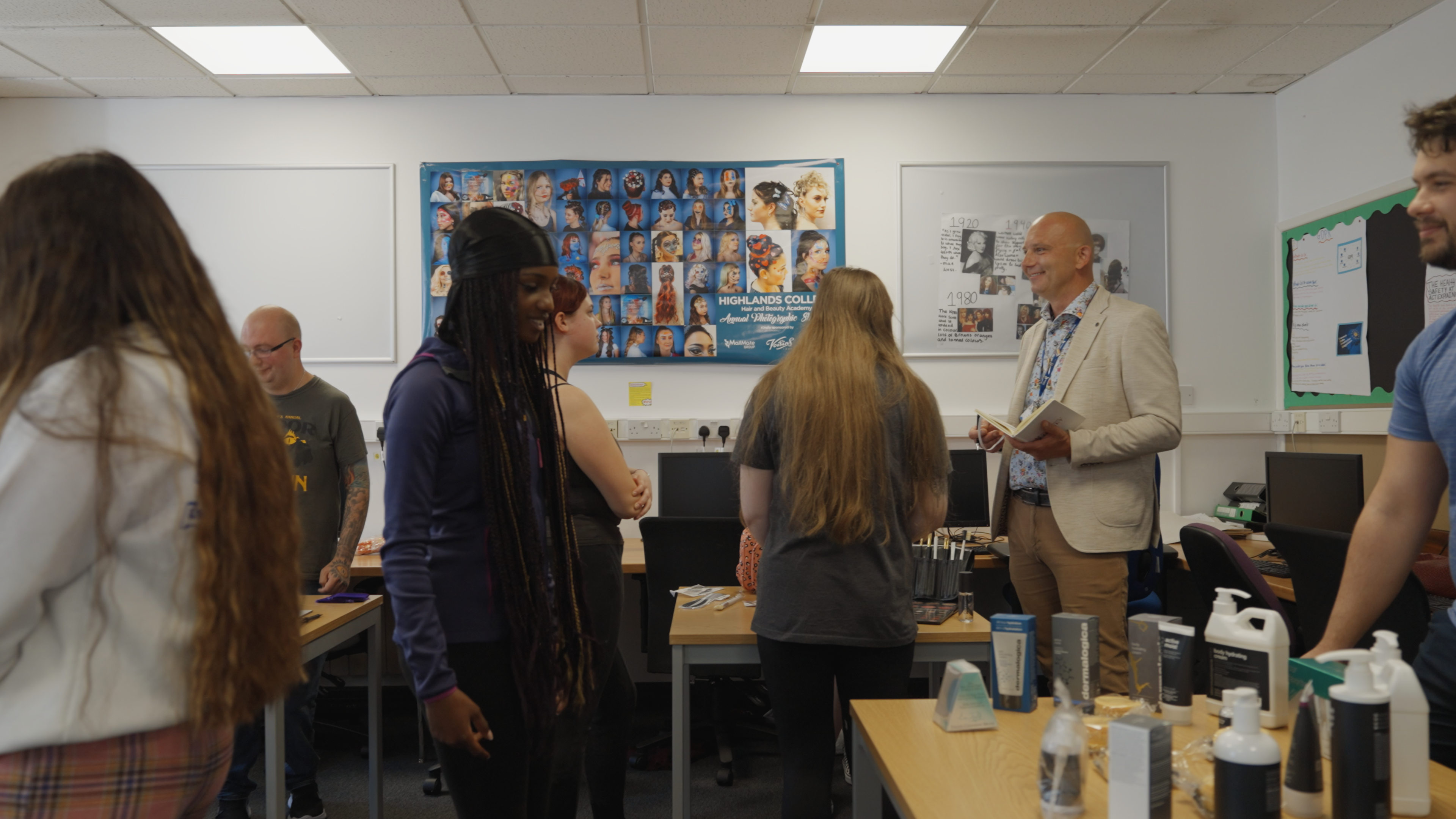A medium shot of a group setting in a classroom. There are tables of various products with people stood behind the tables, smiling.