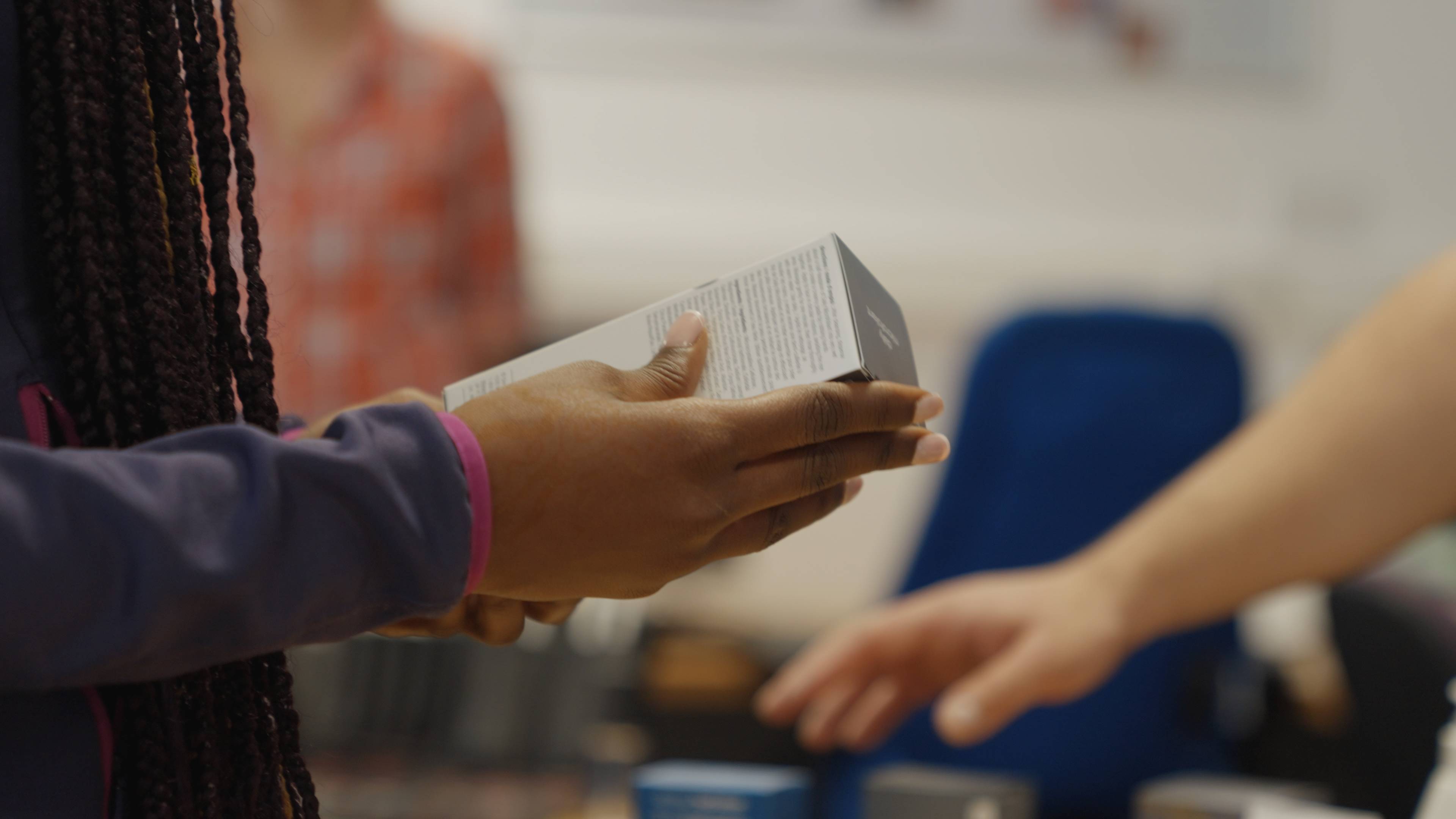 A medium close up of an individual holding a product box in their hands. A blurred hand reaches for another product in the background.