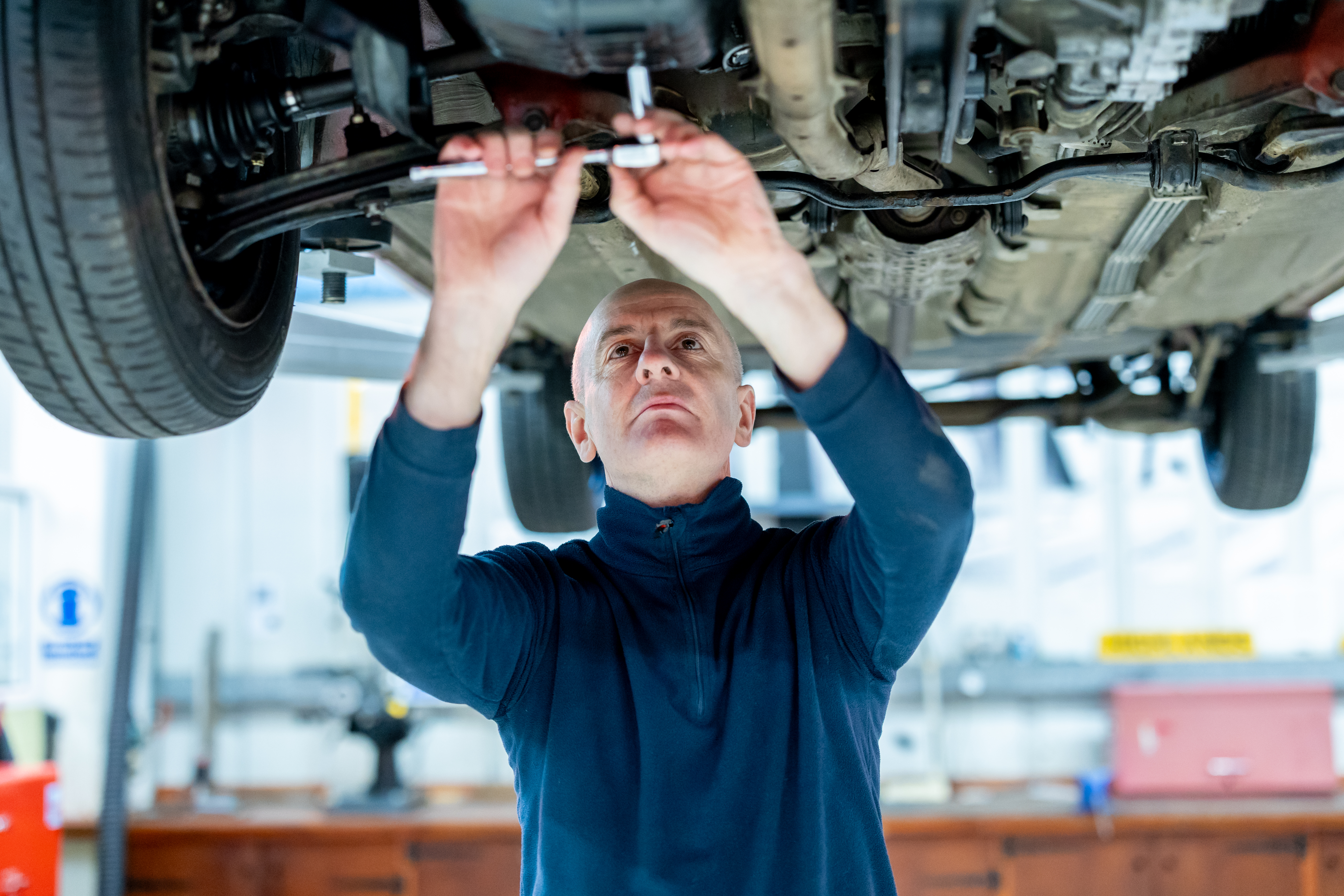 Automotive student at Highlands College working underneath a lifted vehicle in a workshop, using tools to inspect or repair the undercarriage.