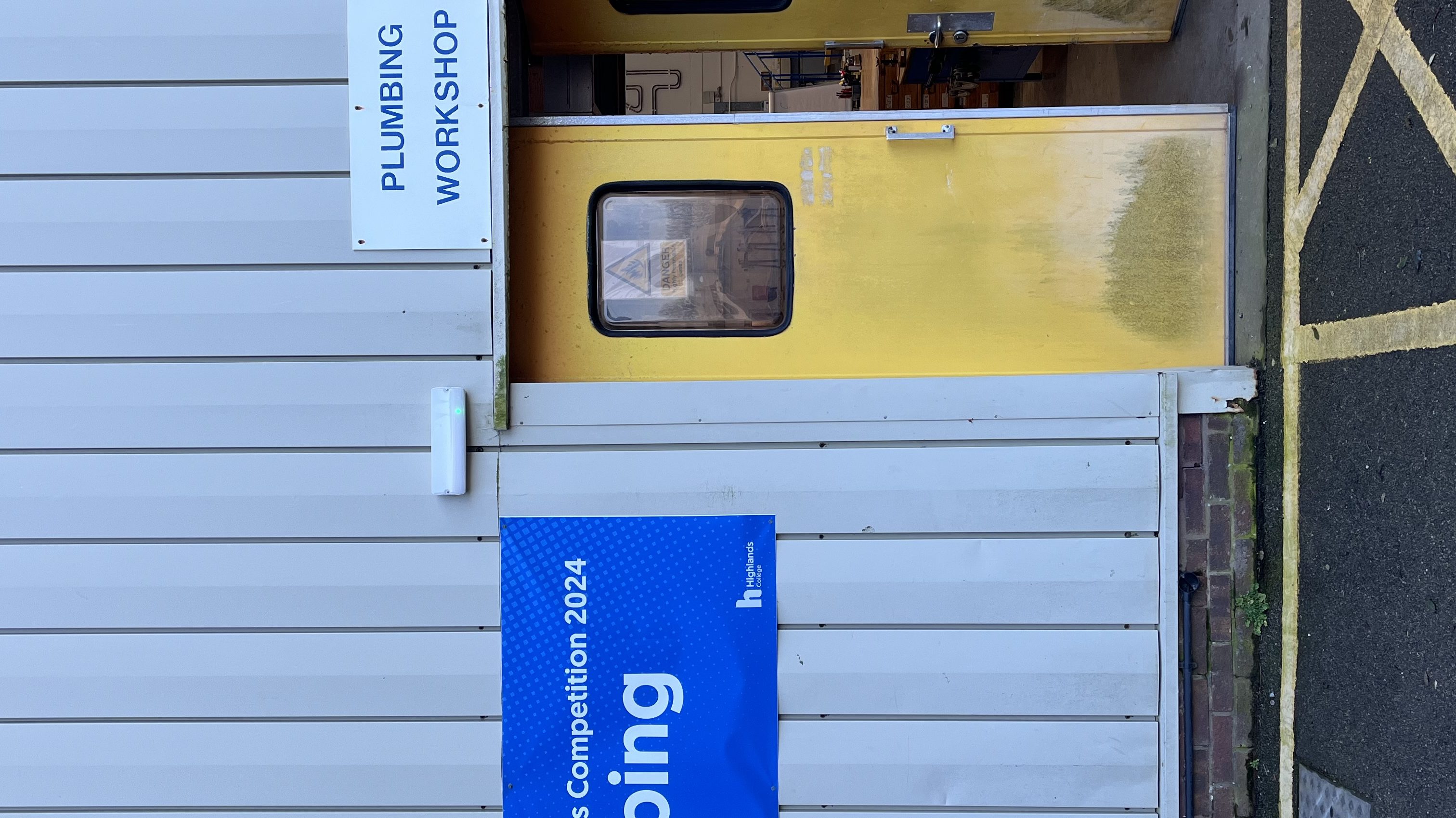 Entrance to a Plumbing Workshop with a yellow door, signage for 'Skills Competition 2024 Plumbing,' and vertical metal siding on the building exterior.