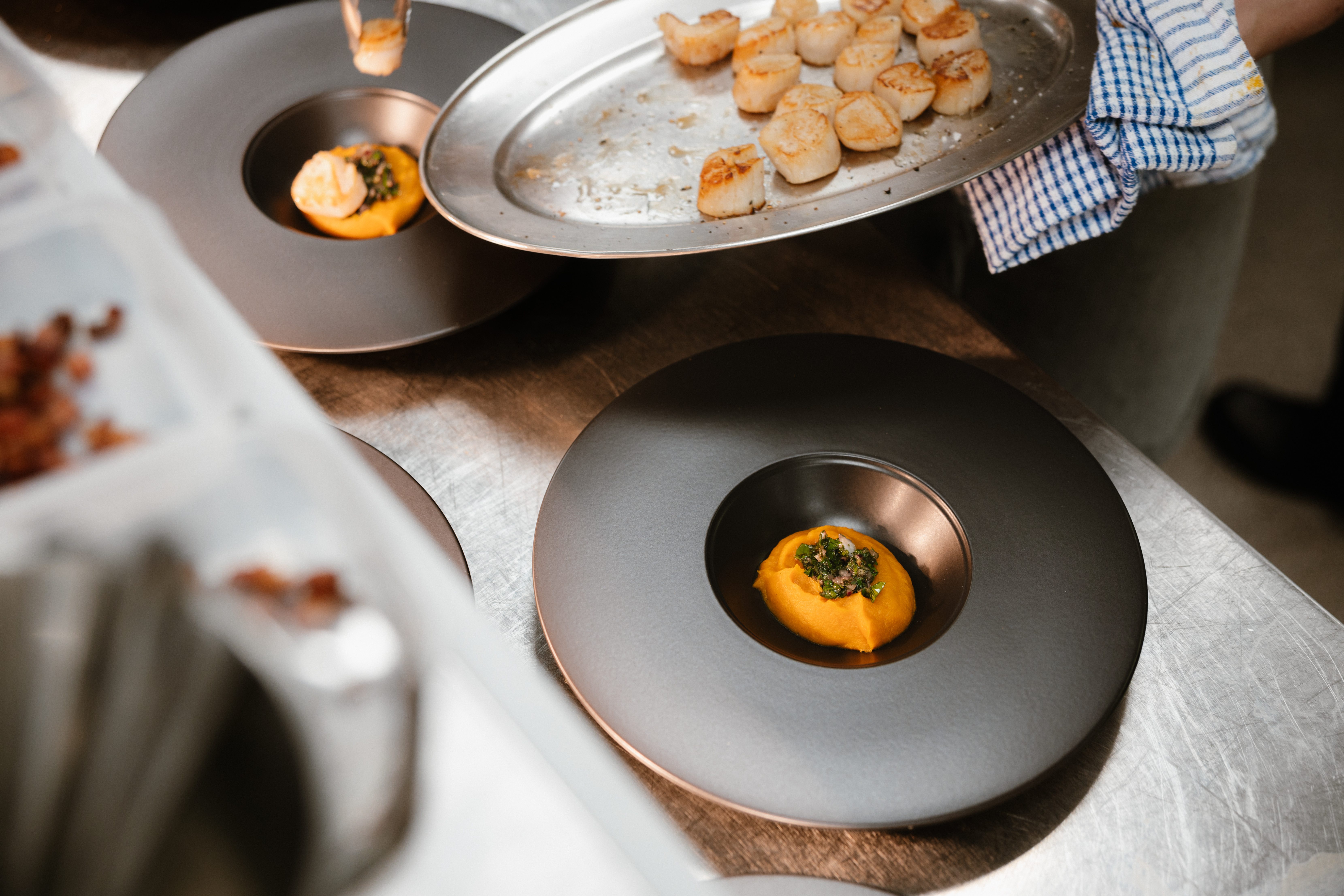 Culinary student at Highlands College plating seared scallops with orange puree and green garnish on black plates in a professional kitchen.