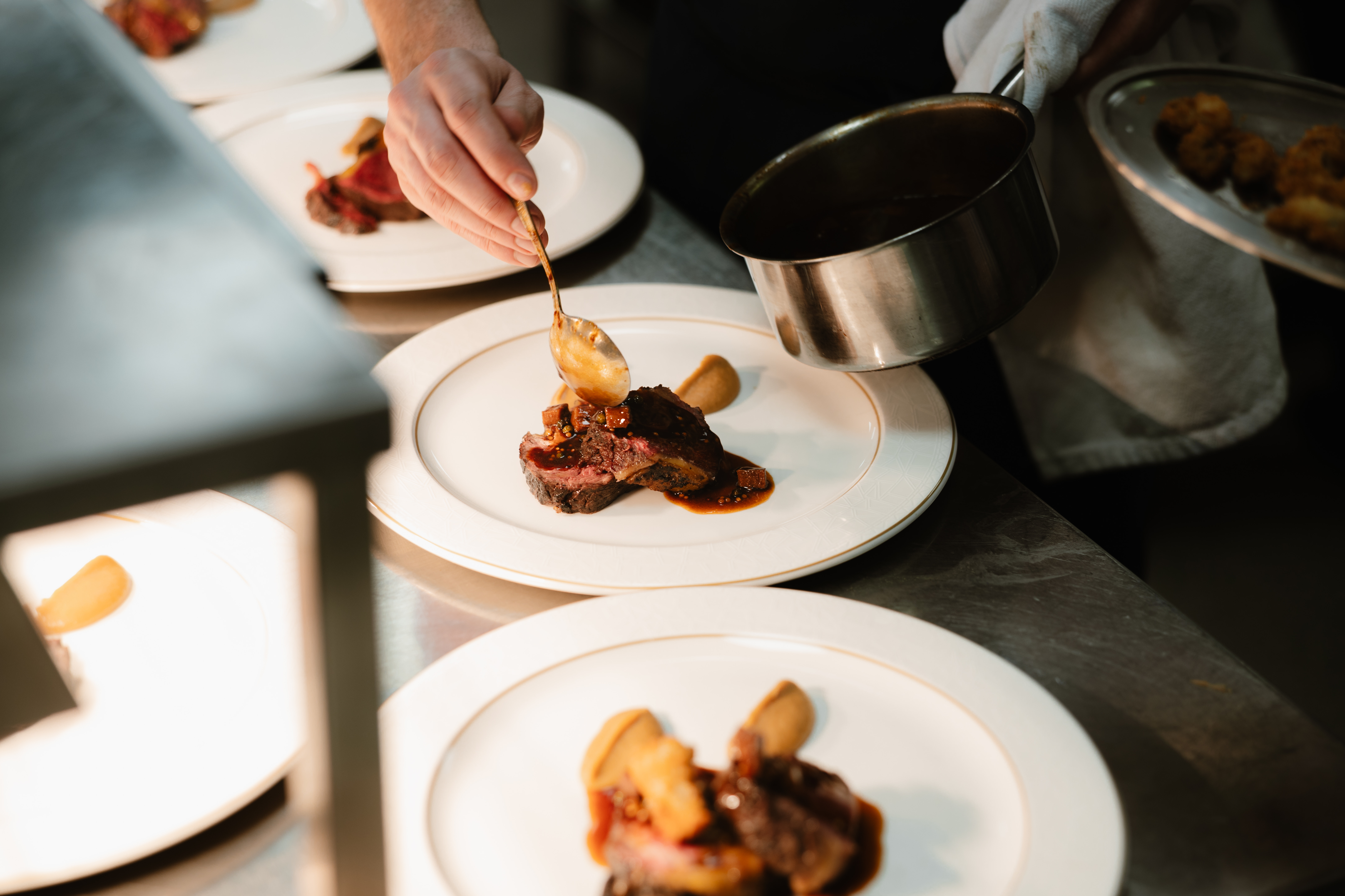 Culinary student at Highlands College plating a meat dish in a professional kitchen, drizzling sauce with a spoon under bright lighting.