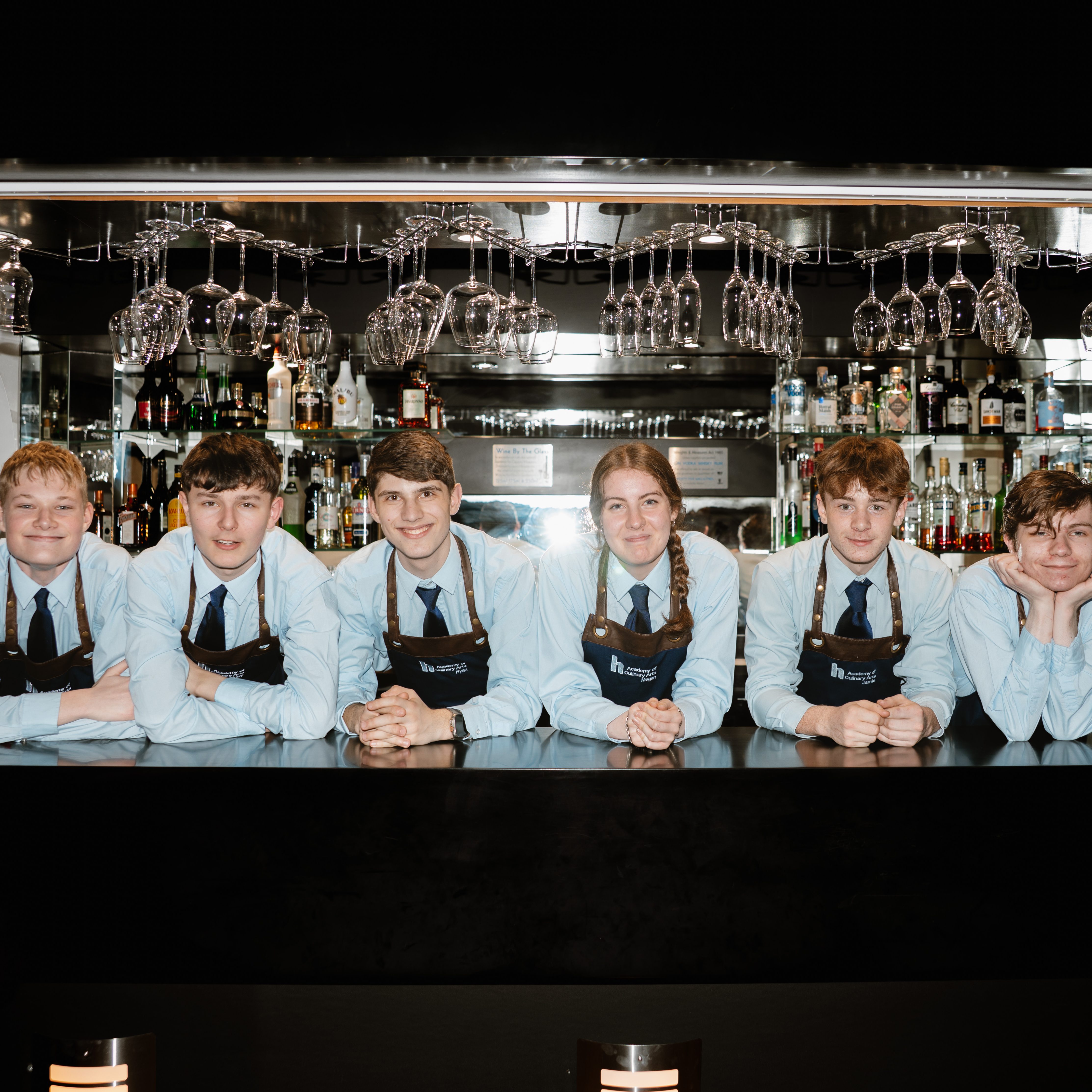 Hospitality students at Highlands College standing behind a bar counter wearing ‘Barista’ aprons, with wine glasses overhead and a stocked beverage shelf in the background.