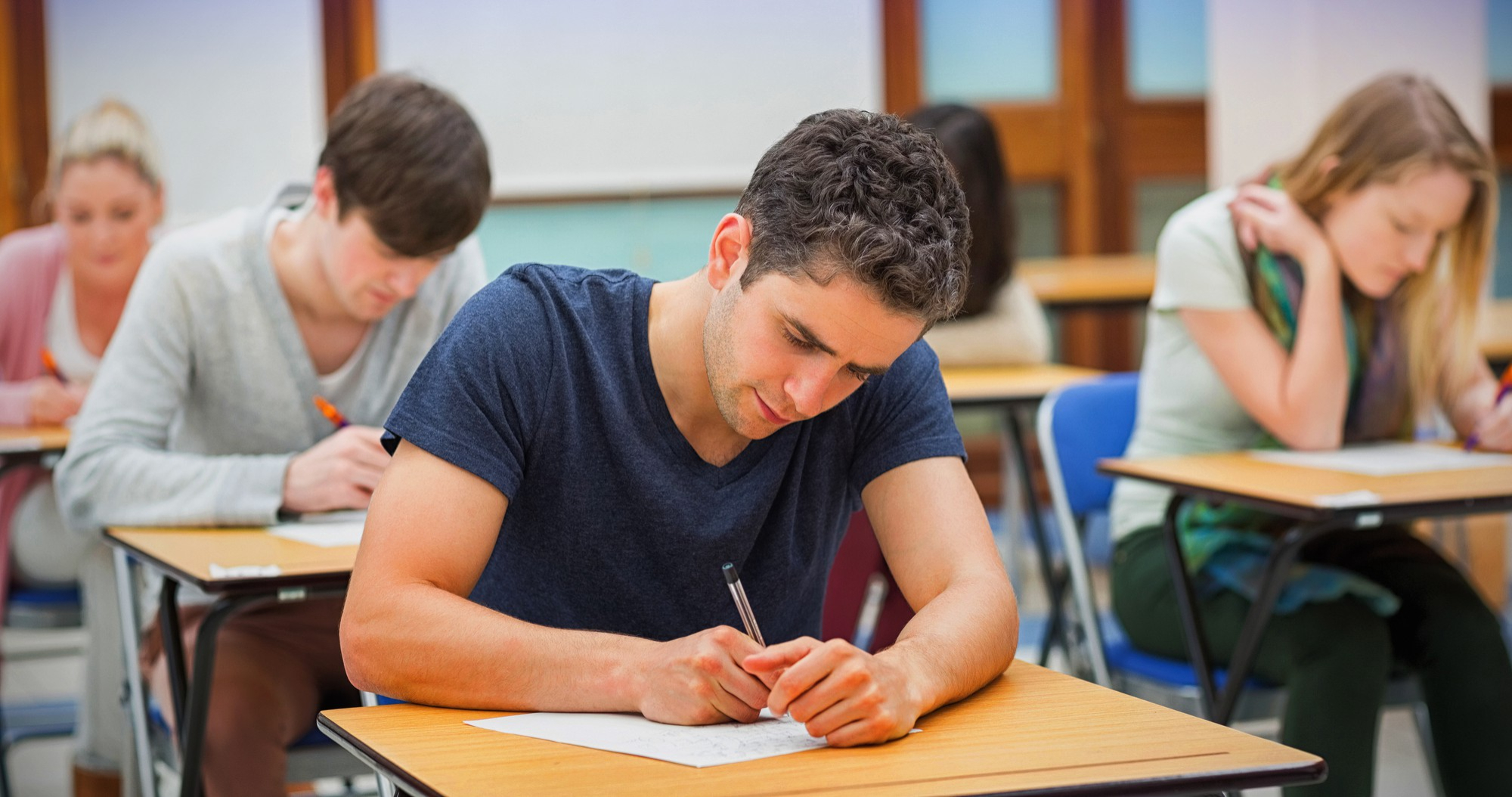 Students seated at wooden desks in a classroom at Highlands College, writing on paper during an exam.