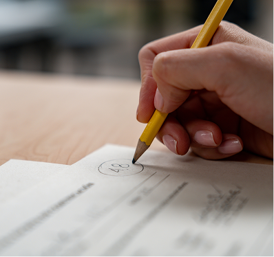 A close-up shot of a light-skinned hand with manicured nails holding a yellow pencil and circling the number 48 printed on a white document or test paper, against the backdrop of a wooden desk.
