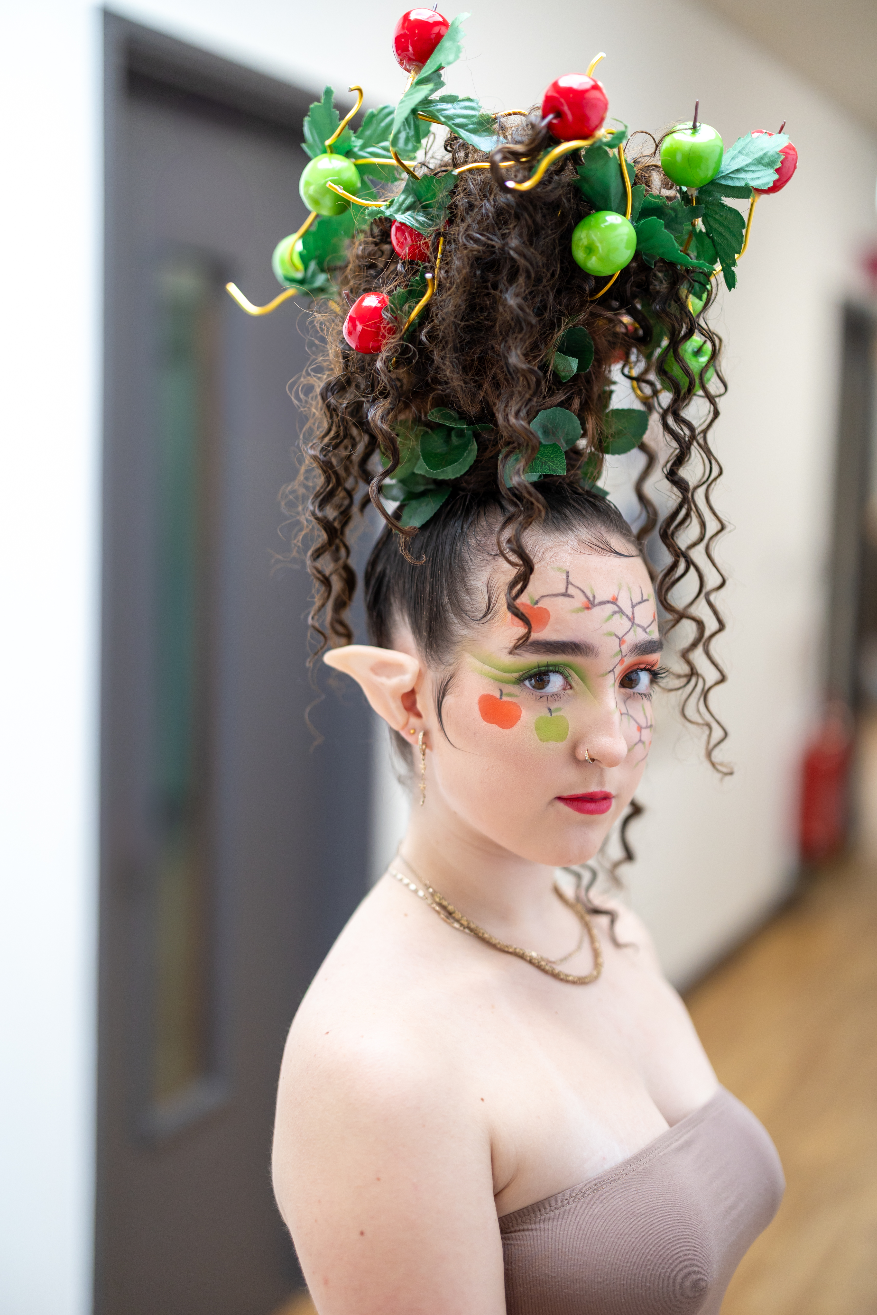 Highlands College student showcasing a creative updo with artificial apples, leaves, and fantasy-themed accessories during a hairdressing styling project.