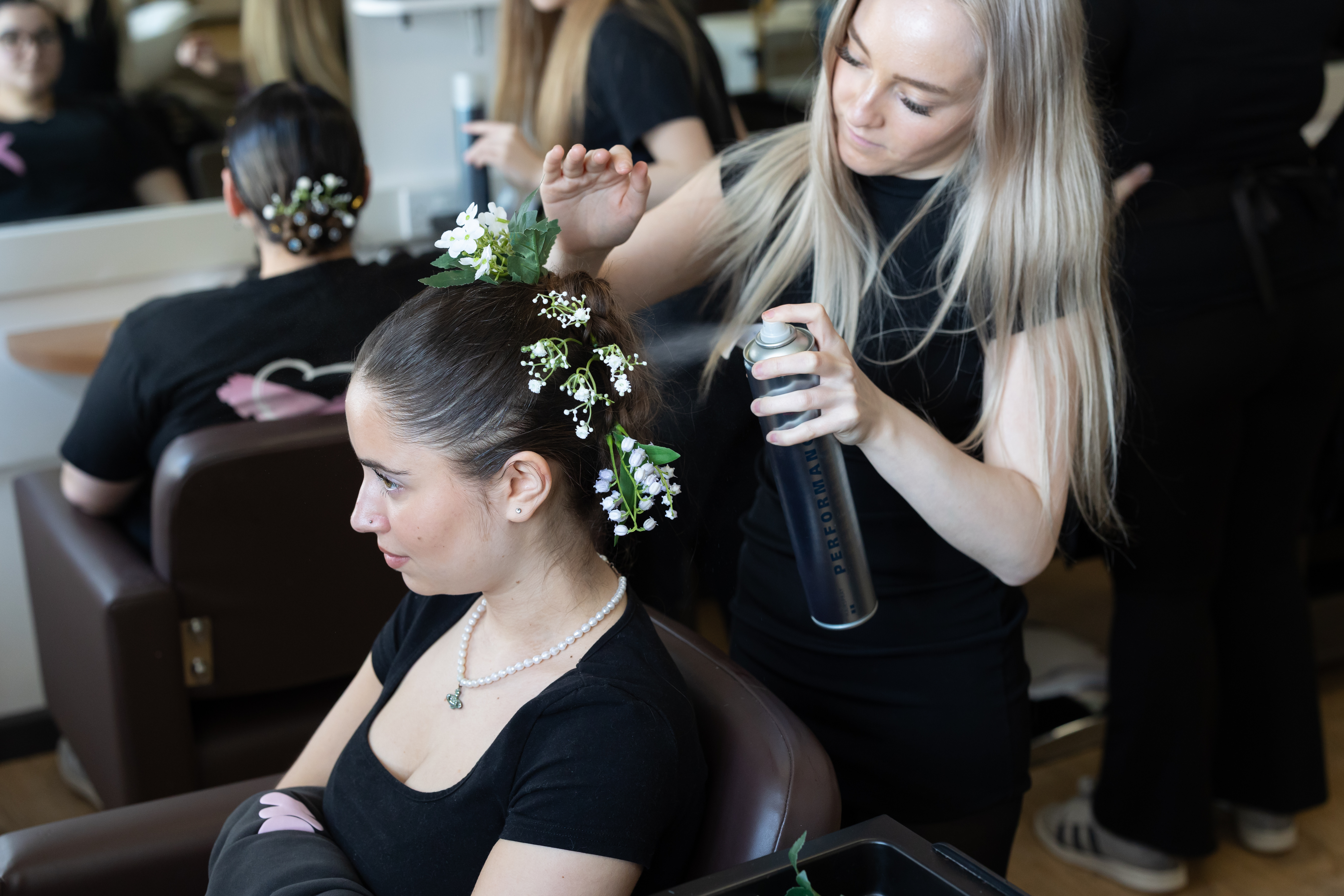 Hairdressing student at Highlands College applying hairspray to a floral bridal hairstyle during a salon training session with multiple clients.