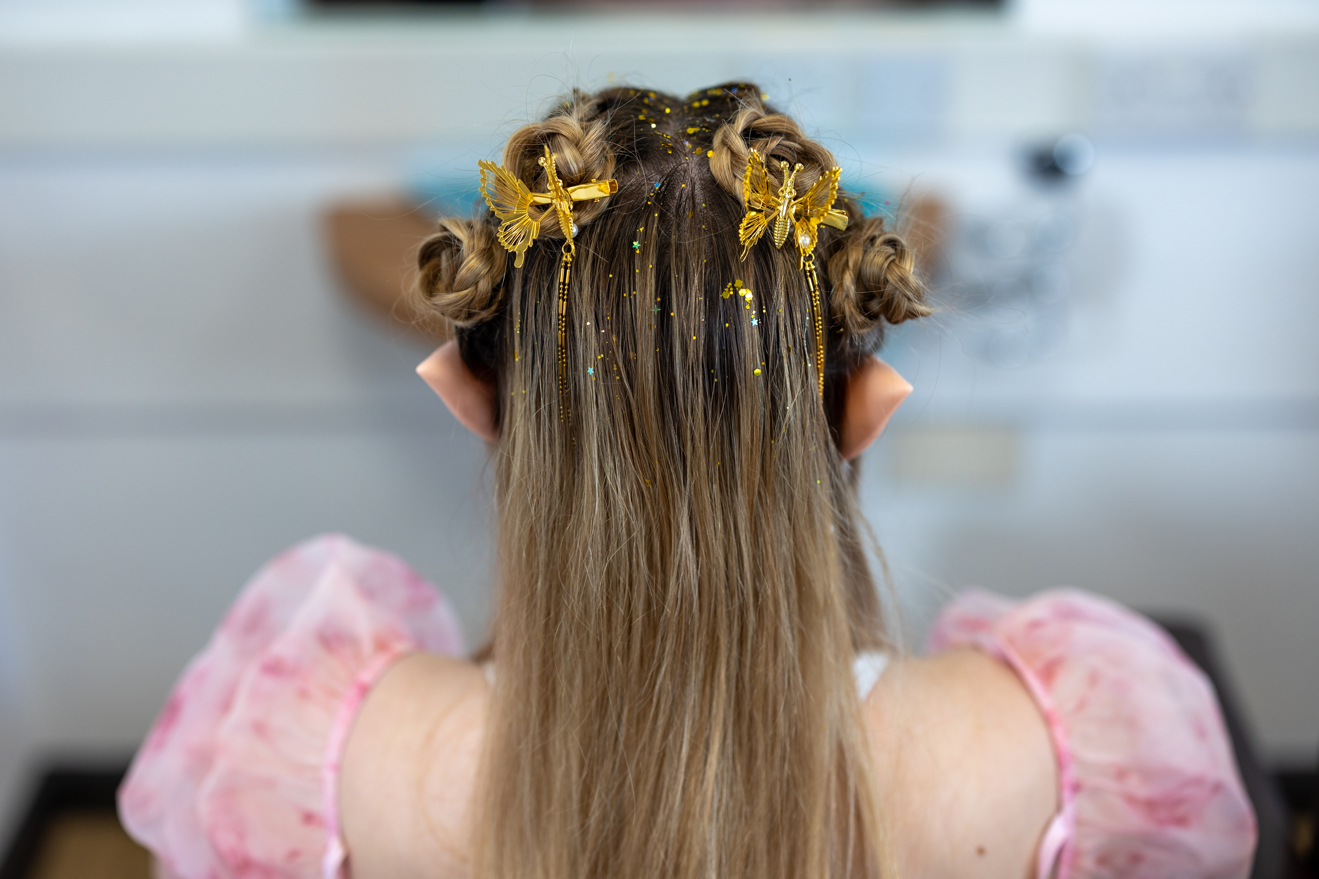 Back view of a person at Highlands College with blonde hair styled in braided buns, decorated with gold butterfly clips and glitter, wearing elf ears and a pink puffy-sleeved outfit.